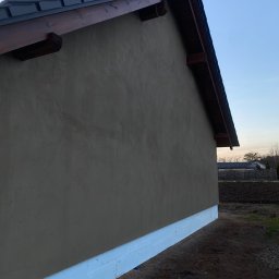 S.EnergyBud Dawid Suduł - Ściana budynku po nałożeniu tynku zewnętrznego, widoczna izolacja fundamentów. Ujęcie z dołu, podkreślające strukturę tynku i detale wykończenia dachu.