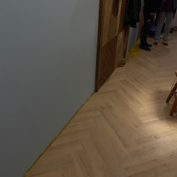 S.EnergyBud Dawid Suduł - Ułożona podłoga z paneli w jodełkę, zabezpieczona taśmą malarską przy ścianie. W tle widoczna drewniana ściana i fragment osób.
