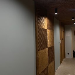 S.EnergyBud Dawid Suduł - Drewniana ściana z dekoracyjnymi panelami w geometryczny wzór, uzupełniona o drewniany sufit z punktowym oświetleniem. W tle widoczne drzwi i fragment korytarza.