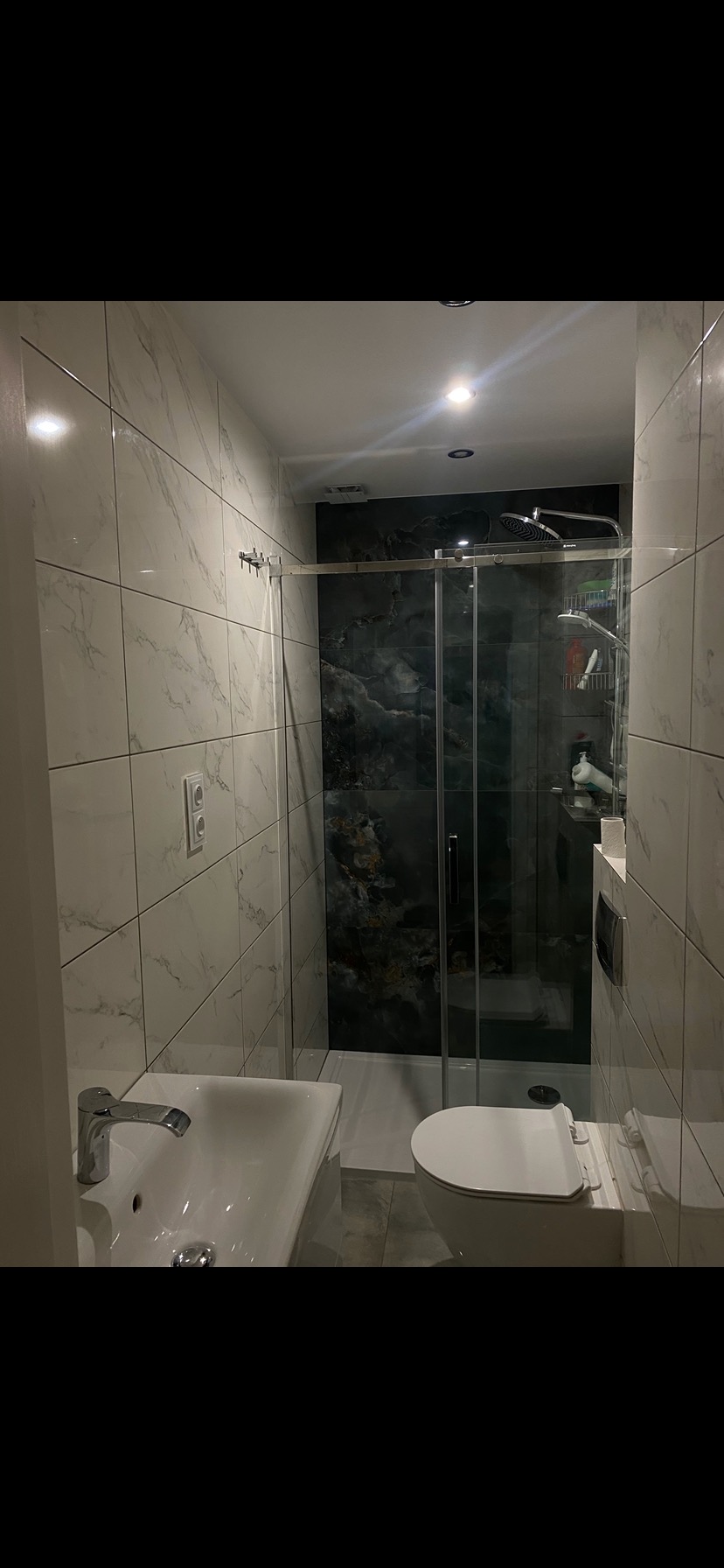 Nowoczesna łazienka z białymi płytkami imitującymi marmur, kabiną prysznicową ze szklanymi drzwiami i toaletą w zabudowie. Czarne akcenty na ścianie pod prysznicem.