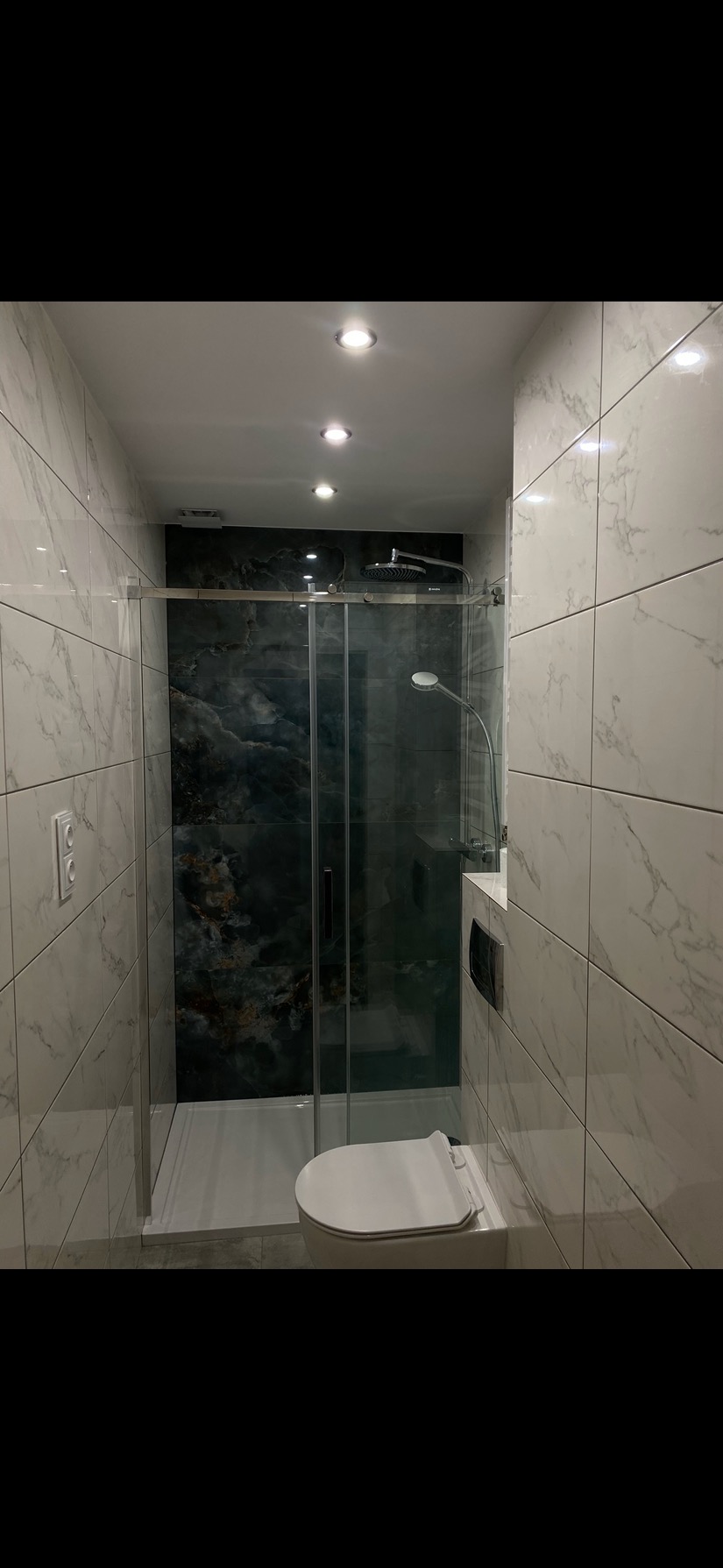Nowoczesna łazienka z kabiną prysznicową typu walk-in, marmurowymi płytkami i podwieszaną toaletą. Czarne akcenty na ścianie prysznica i suficie.