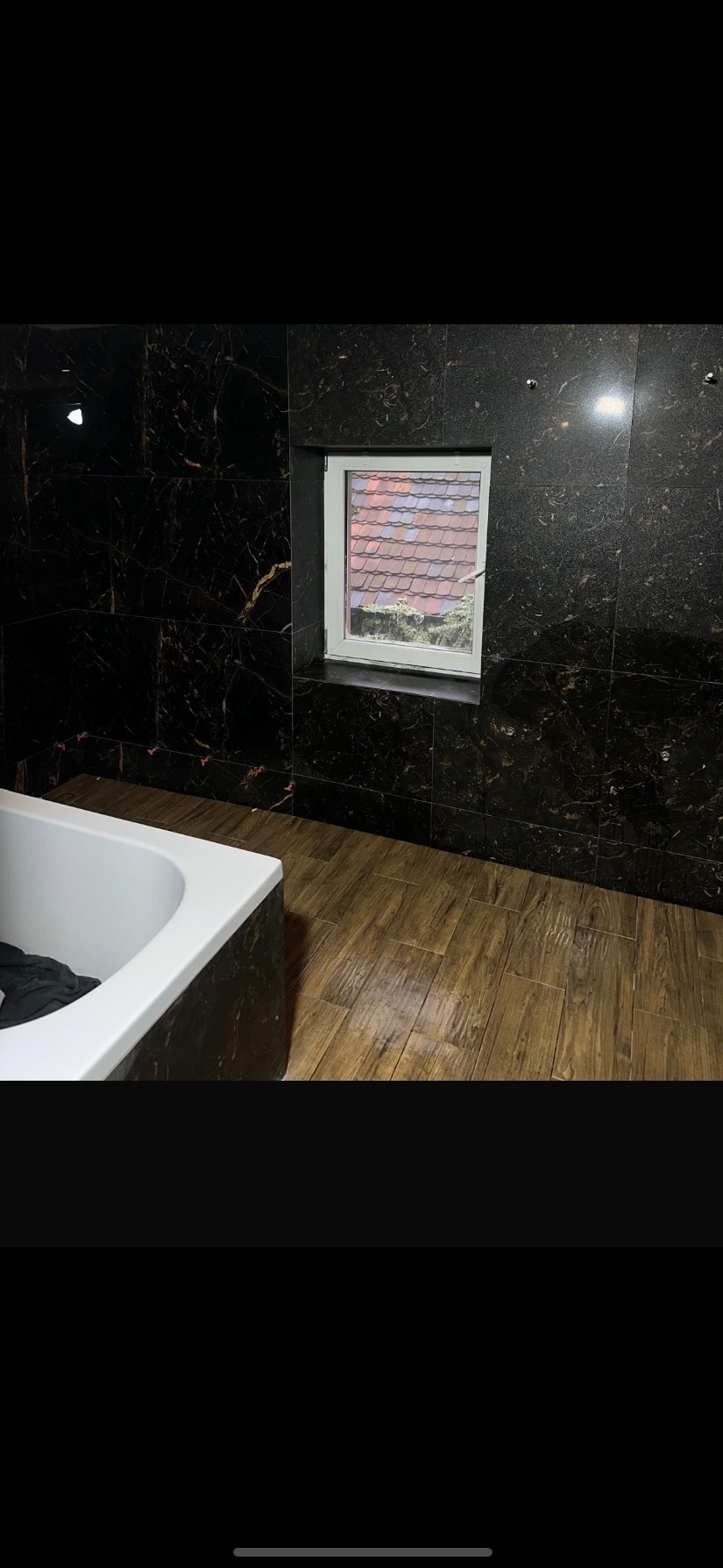 Wykończona łazienka z wanną, okienkiem i ciemnymi płytkami imitującymi marmur na ścianach i podłodze. Widok z góry na dach budynku za oknem.