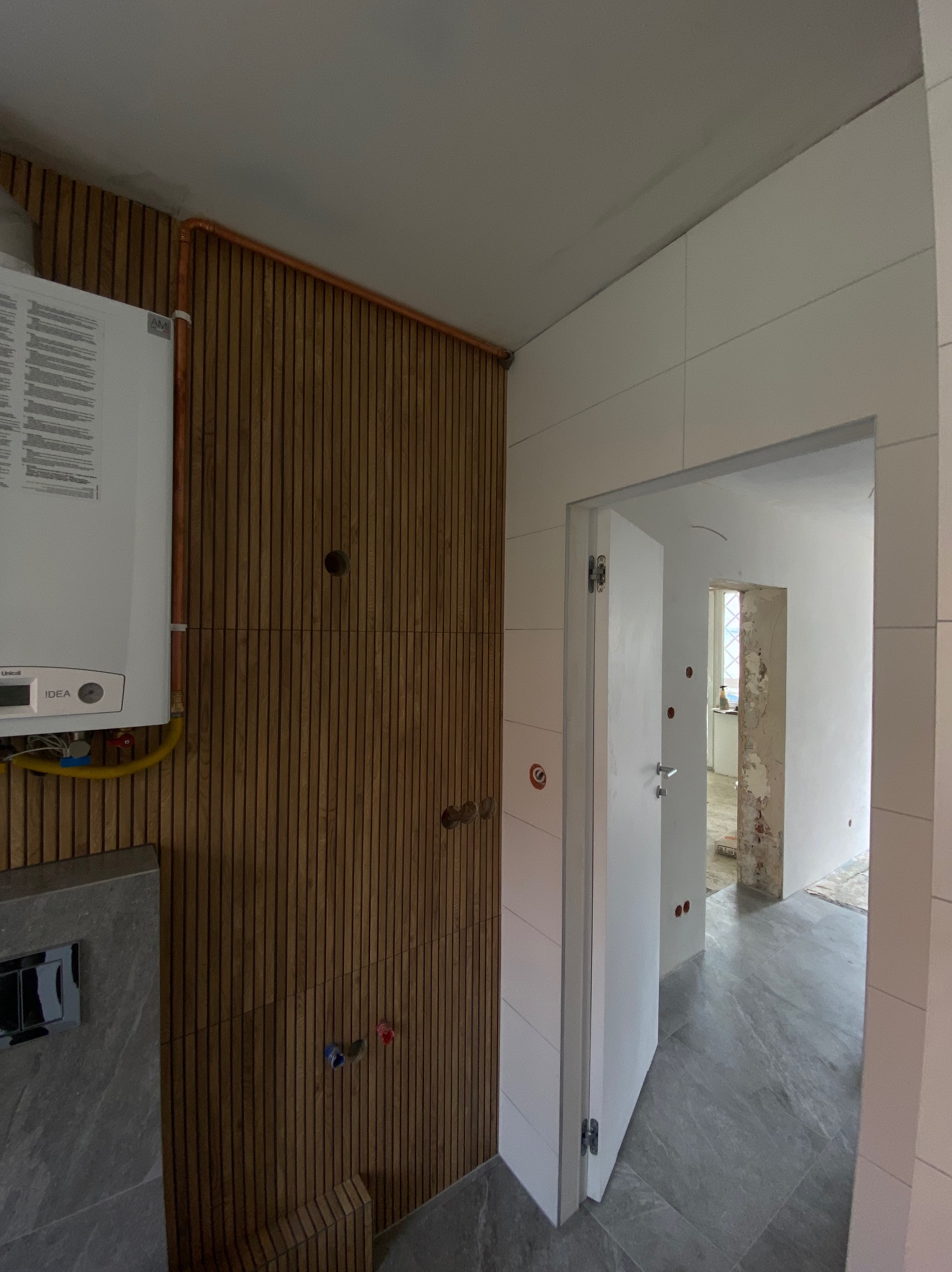 Nowoczesna łazienka w trakcie wykończenia: drewniana boazeria, marmurowe płytki podłogowe, piec gazowy i otwarte drzwi do remontowanego pomieszczenia.