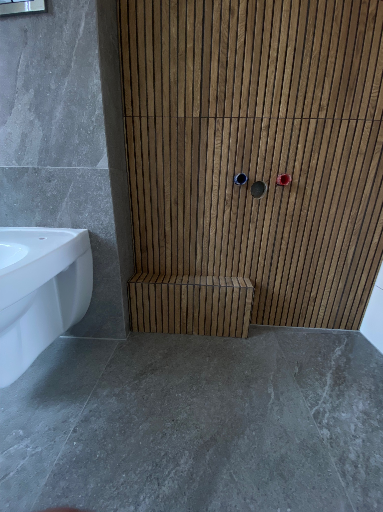 Nowoczesna łazienka: szara płytka, drewniane lamele na ścianie z widocznymi przyłączami wodnymi i biała umywalka. Minimalistyczny design.