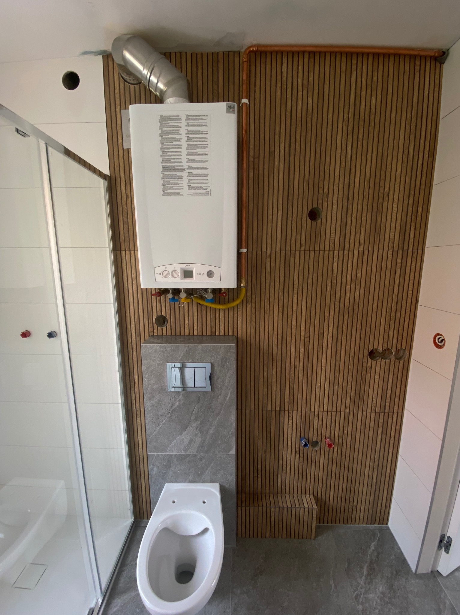 Nowoczesna łazienka z kabiną prysznicową, wiszącą toaletą i piecem gazowym na ścianie z drewnianymi lamelami. Widoczne miedziane rury i szara płytka podtynkowa.