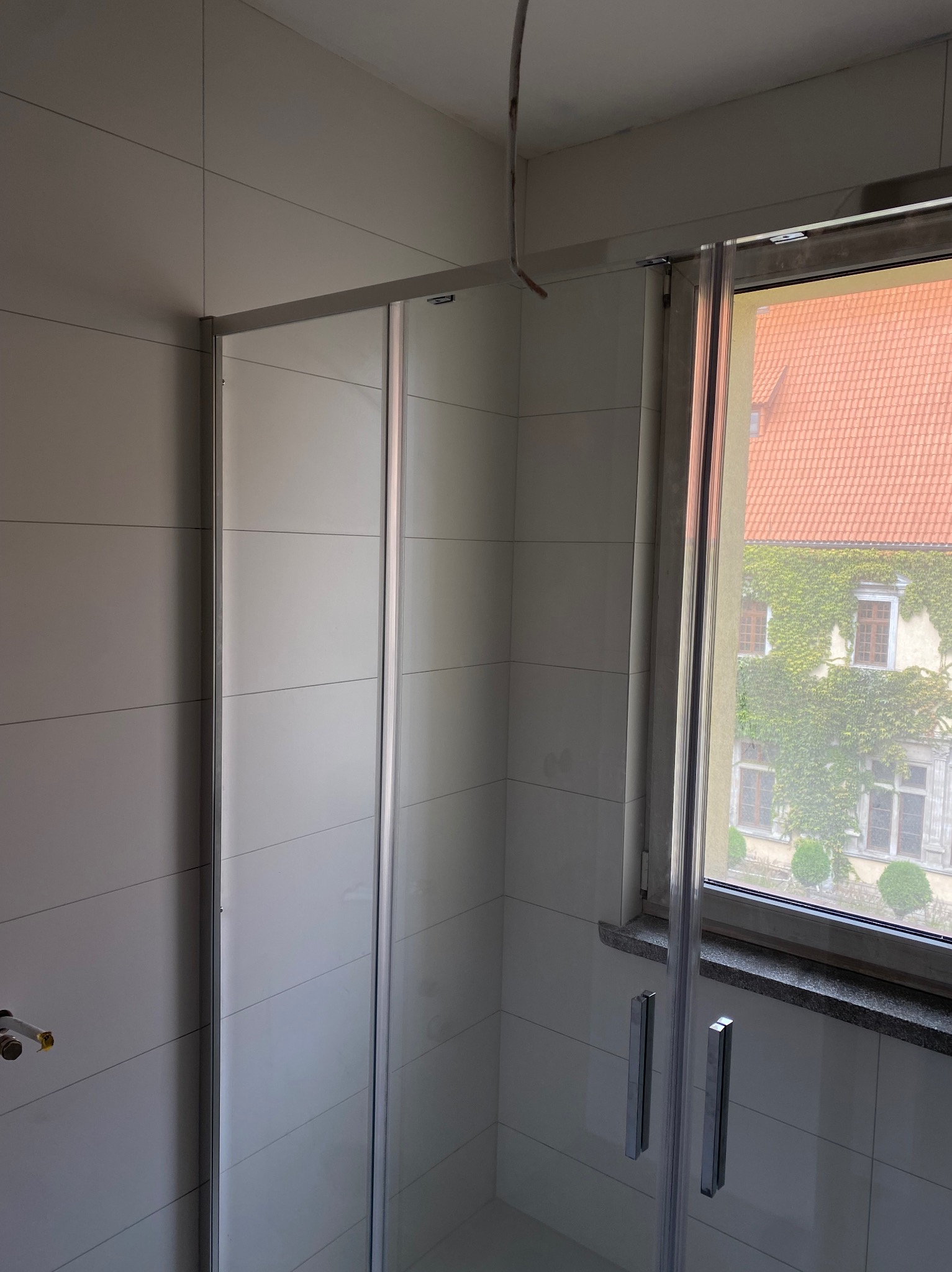Wykończona łazienka z kabiną prysznicową, jasnymi płytkami i oknem z widokiem na budynek porośnięty bluszczem. Widoczne niedokończone podłączenie kabla.