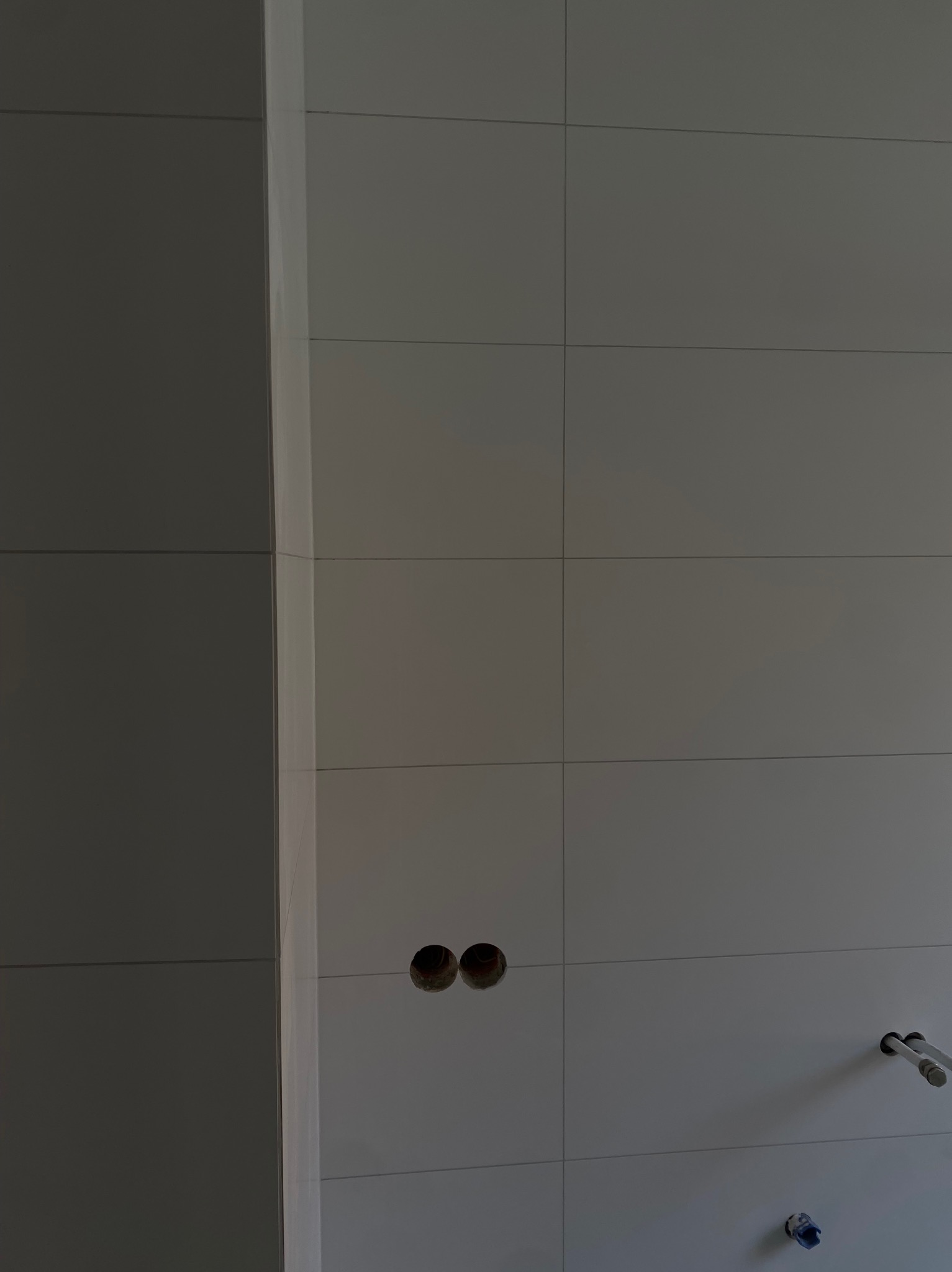 Ściana wyłożona jasnymi, kwadratowymi płytkami z widocznymi otworami na instalacje i fragmentem rurki, ukazująca etap prac wykończeniowych w łazience.