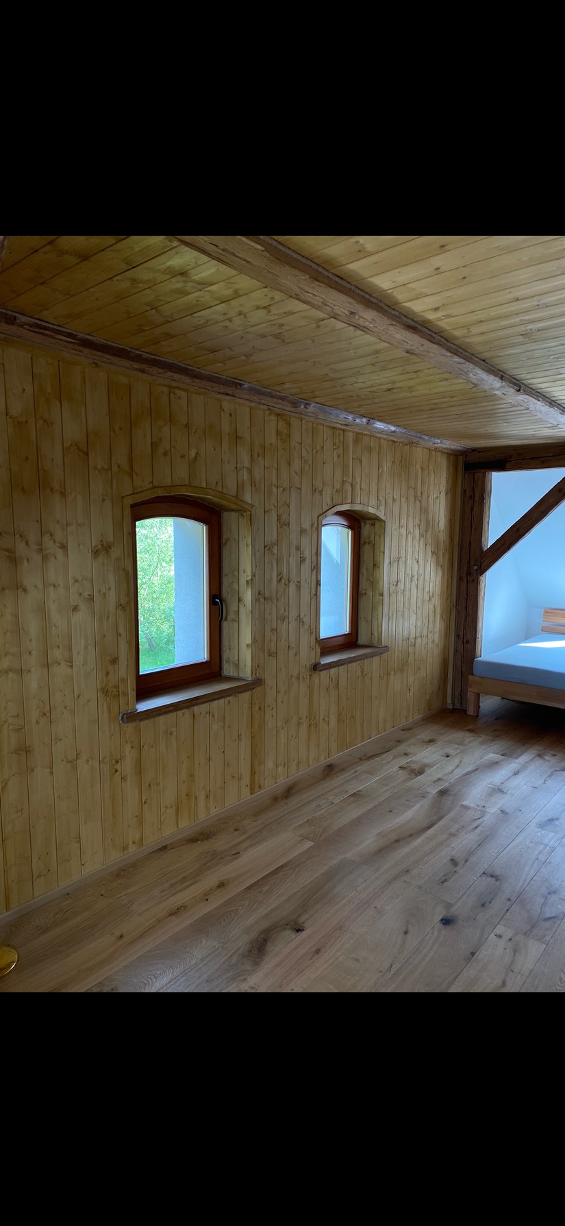 Drewniane wykończenie wnętrza: boazeria ścienna, podłoga z desek, drewniany strop i okna z zaokrąglonymi ramami. W tle widoczne łóżko.