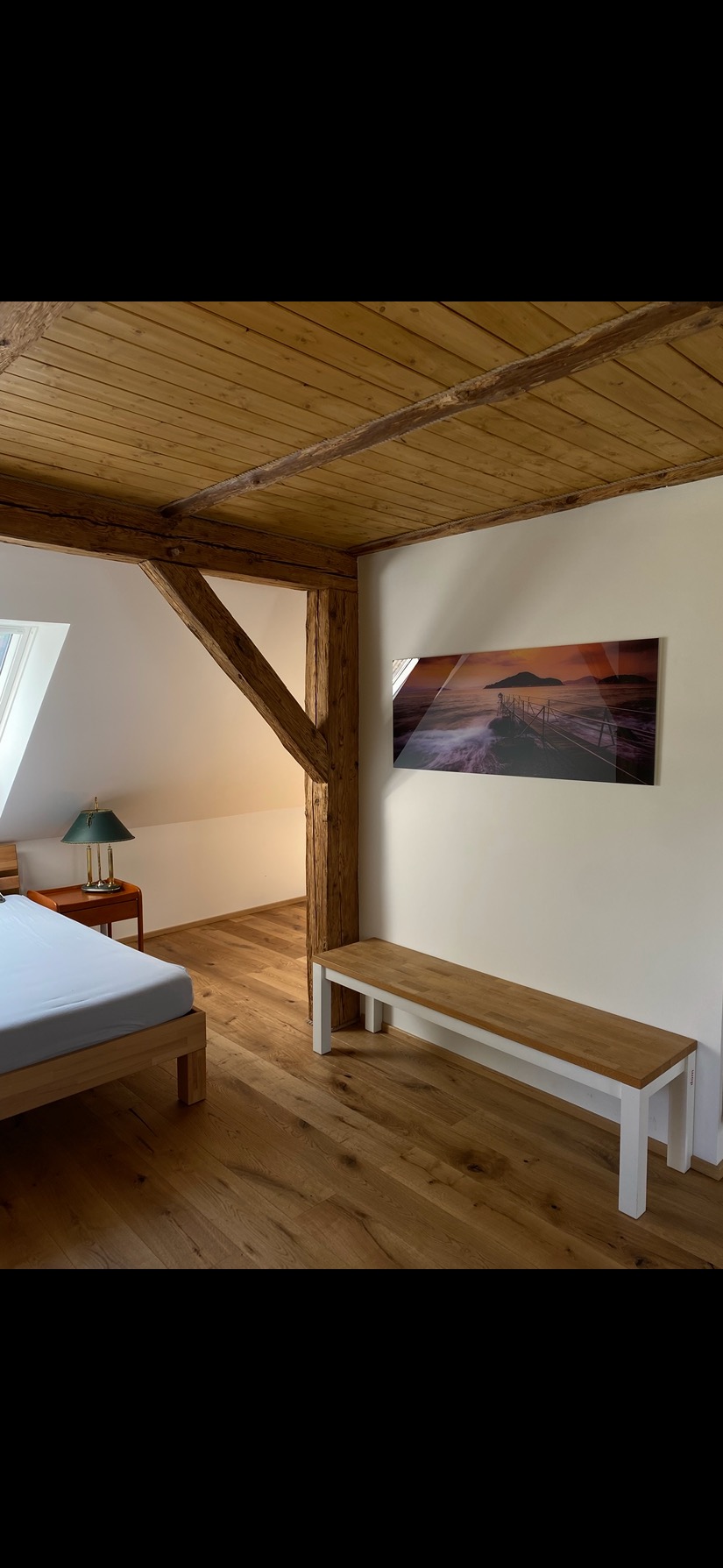 Drewniana podłoga w pokoju na poddaszu z widoczną belką konstrukcyjną, łóżkiem i obrazem z motywem morskim na ścianie.