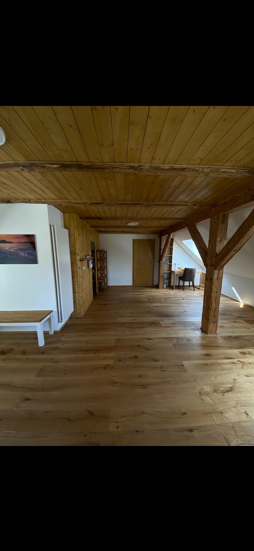 Jasne panele podłogowe w przestronnym pokoju z drewnianym sufitem i filarami. Widoczne grzejnik, obraz na ścianie i elementy wyposażenia wnętrza.