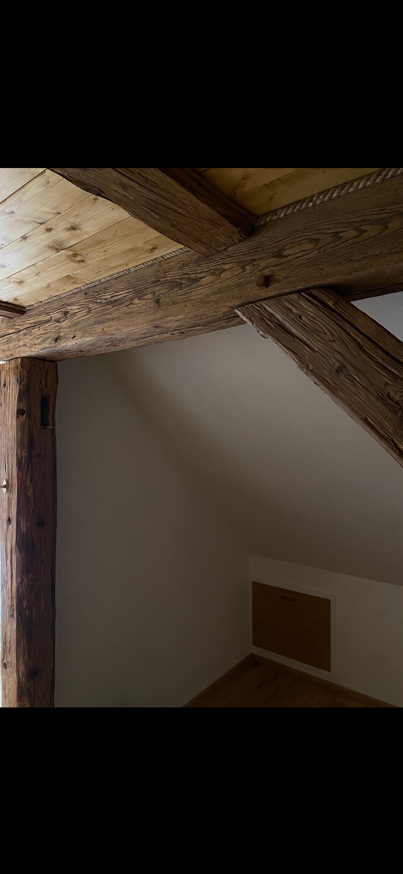 Drewniane belki stropowe i kolumna w jasnym wnętrzu poddasza, widoczne detale tekstury drewna i subtelne oświetlenie.