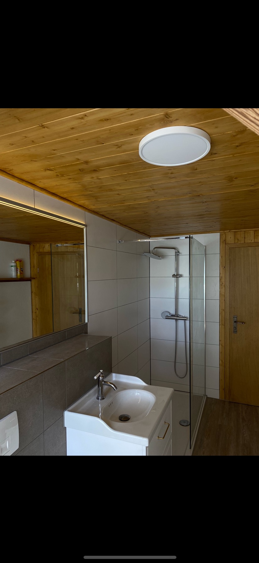 Jasna łazienka z drewnianym sufitem: kabina prysznicowa za szklaną ścianką, biała umywalka z szafką, lustro z podświetleniem LED. Minimalistyczny design.