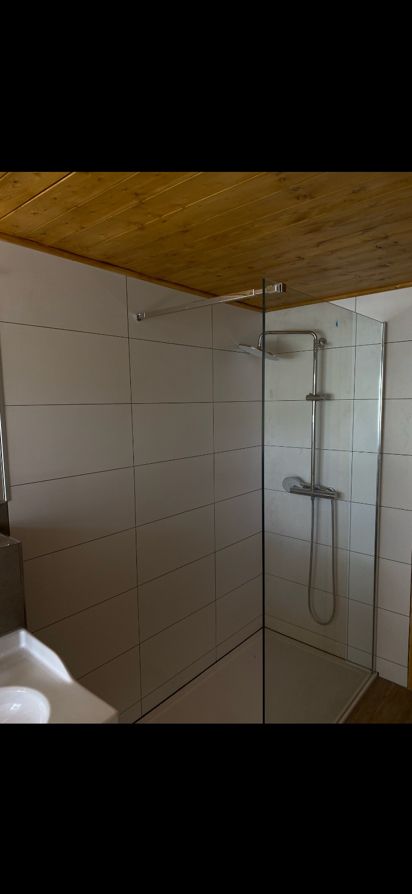 Wykończona kabina prysznicowa z białymi płytkami i drewnianym sufitem. Nowoczesna armatura, minimalistyczny design. Widok na kabinę z boku, z elementami umywalki.