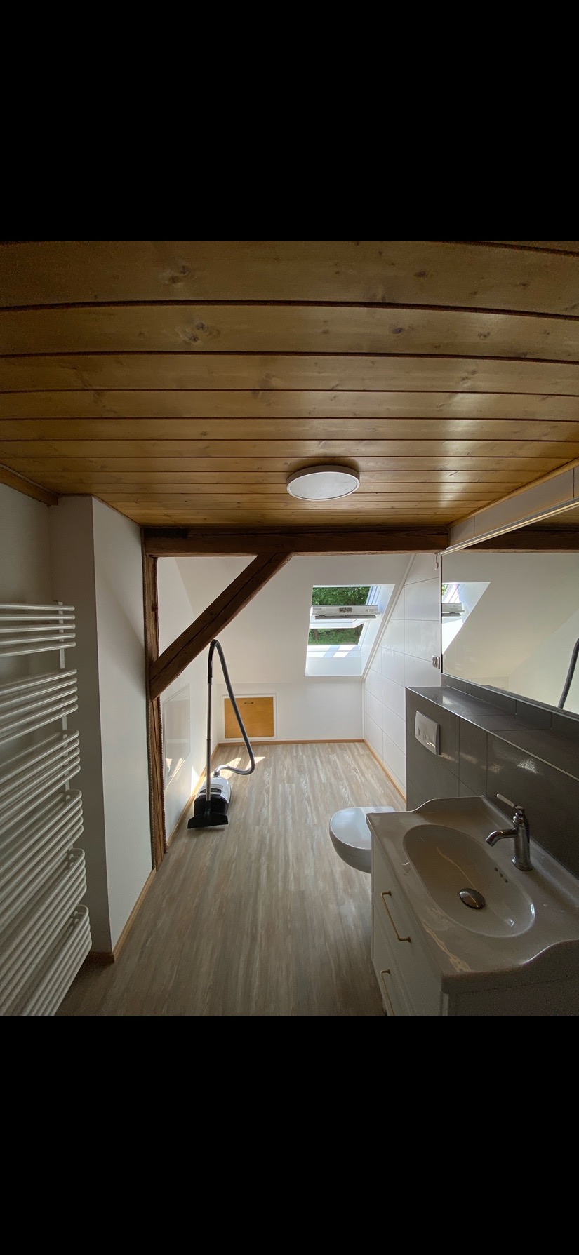 Jasna łazienka na poddaszu z drewnianym stropem, umywalką z szafką, grzejnikiem i odkurzaczem. Okno dachowe wpuszcza naturalne światło.
