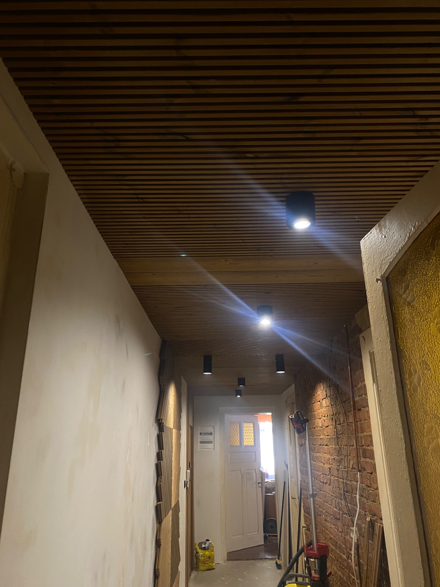Korytarz w trakcie remontu: drewniany sufit lamelowy z oświetleniem punktowym, ściany częściowo wykończone, fragment odsłoniętej cegły.