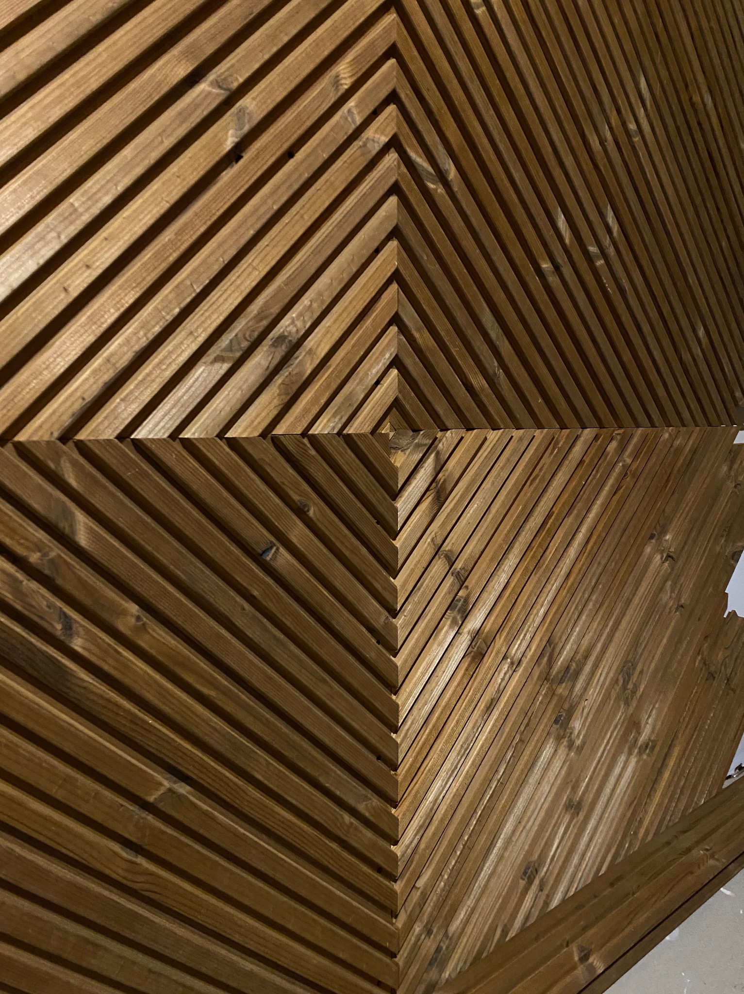 Drewniana ściana z listew ułożonych w jodełkę, tworząca geometryczny wzór. Listwy w ciepłym, brązowym odcieniu z widocznym usłojeniem.