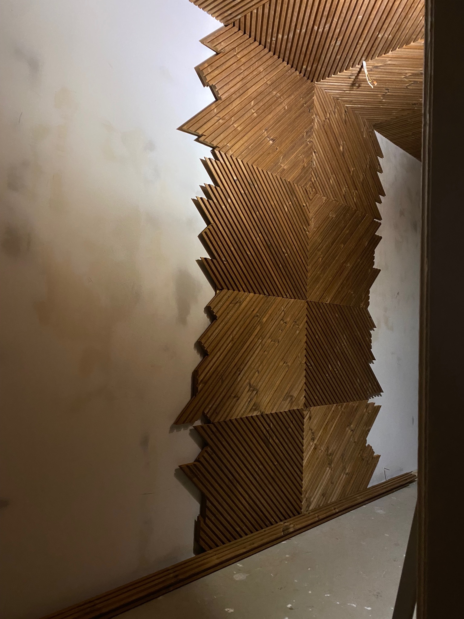 Drewniana mozaika ścienna w trakcie montażu. Ukończony fragment z desek ułożonych pod kątem, surowa ściana obok. Widoczne listwy przypodłogowe.