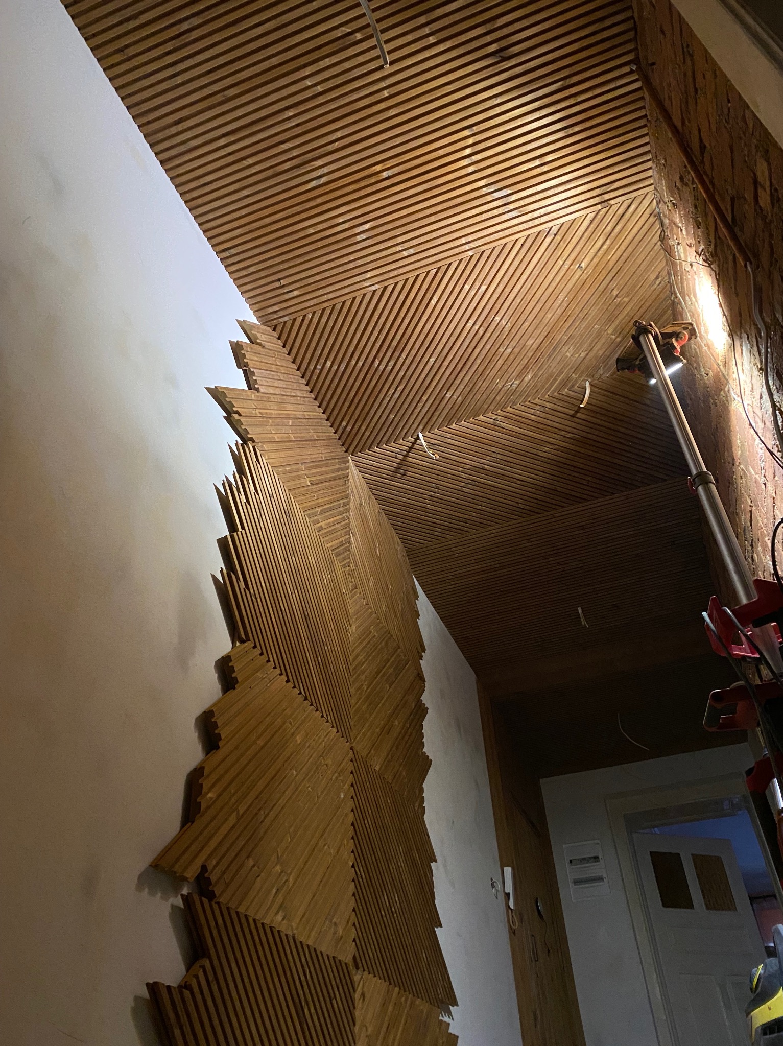 Drewniana boazeria ułożona we wzór na ścianie i suficie, widok na korytarz z drzwiami. Montaż oświetlenia na ścianie z cegły. Nieregularny kształt drewnianej dekoracji.