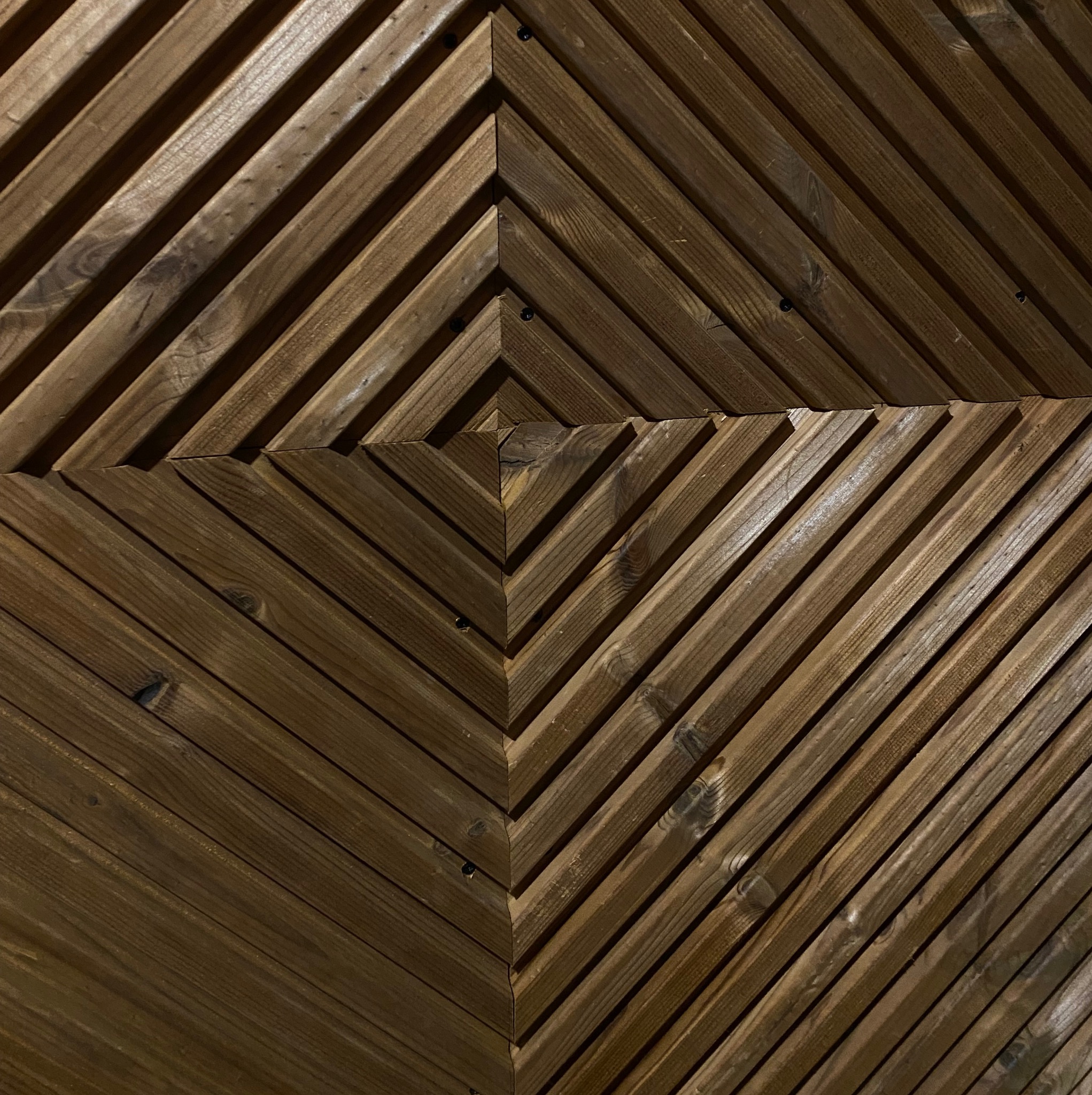 Drewniana ściana z desek ułożonych w geometryczny wzór rombów, z widocznymi śrubami mocującymi, w ciepłej tonacji brązu.