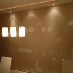 Przygotowanie ściany do malowania: szpachlowana ściana w odcieniach beżu z widocznymi łatami szpachli, narzędzia do szlifowania na stole, wiszące lampy z białymi kloszami i grzejnik.