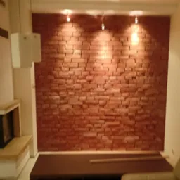 Ściana z cegieł dekoracyjnych podświetlona trzema lampami punktowymi, fragment kominka i sofy w tle, widok na wykończone wnętrze salonu.