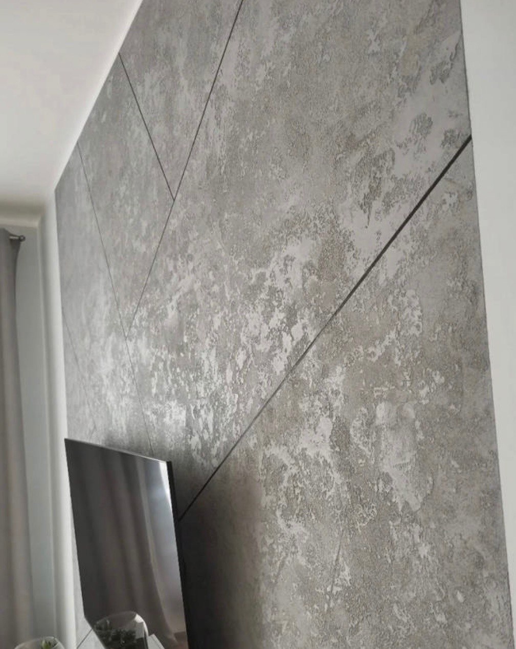 Dekoracyjna ściana wewnętrzna z szarą, strukturalną powłoką imitującą beton, podzielona czarnymi listwami na geometryczne panele w kształcie strzałek.