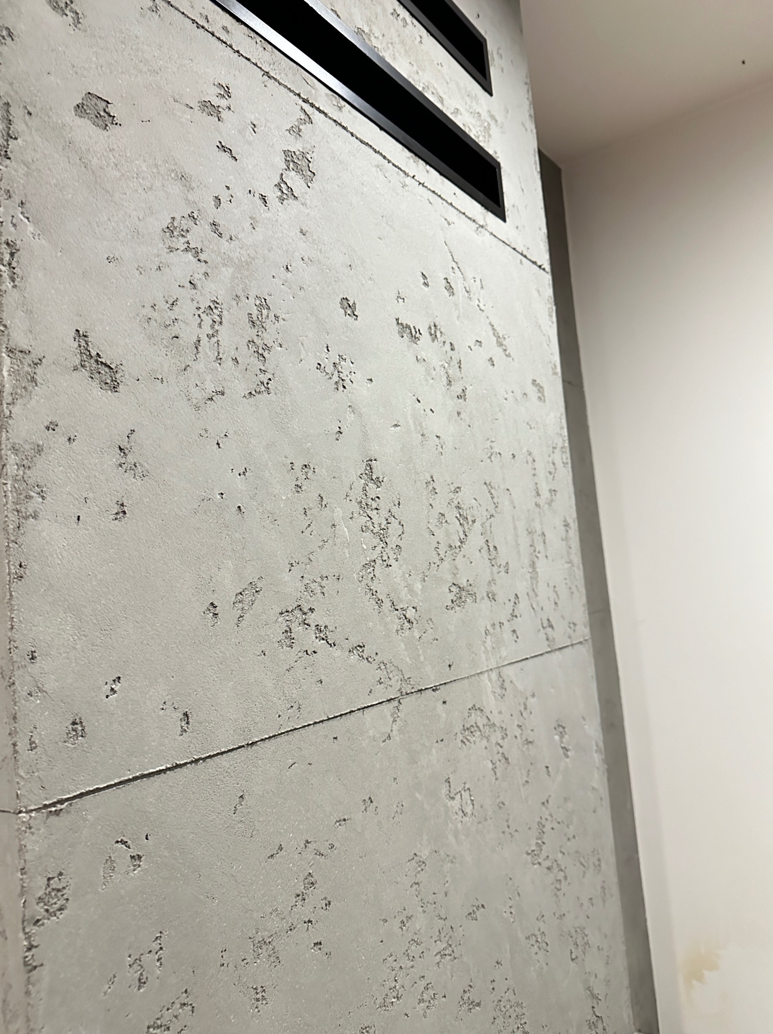 Ściana wewnętrzna wykończona szarym betonem architektonicznym z widocznymi porami i fakturą, fragment sufitu z czarnymi kratkami wentylacyjnymi, ujęcie z dołu pod kątem.