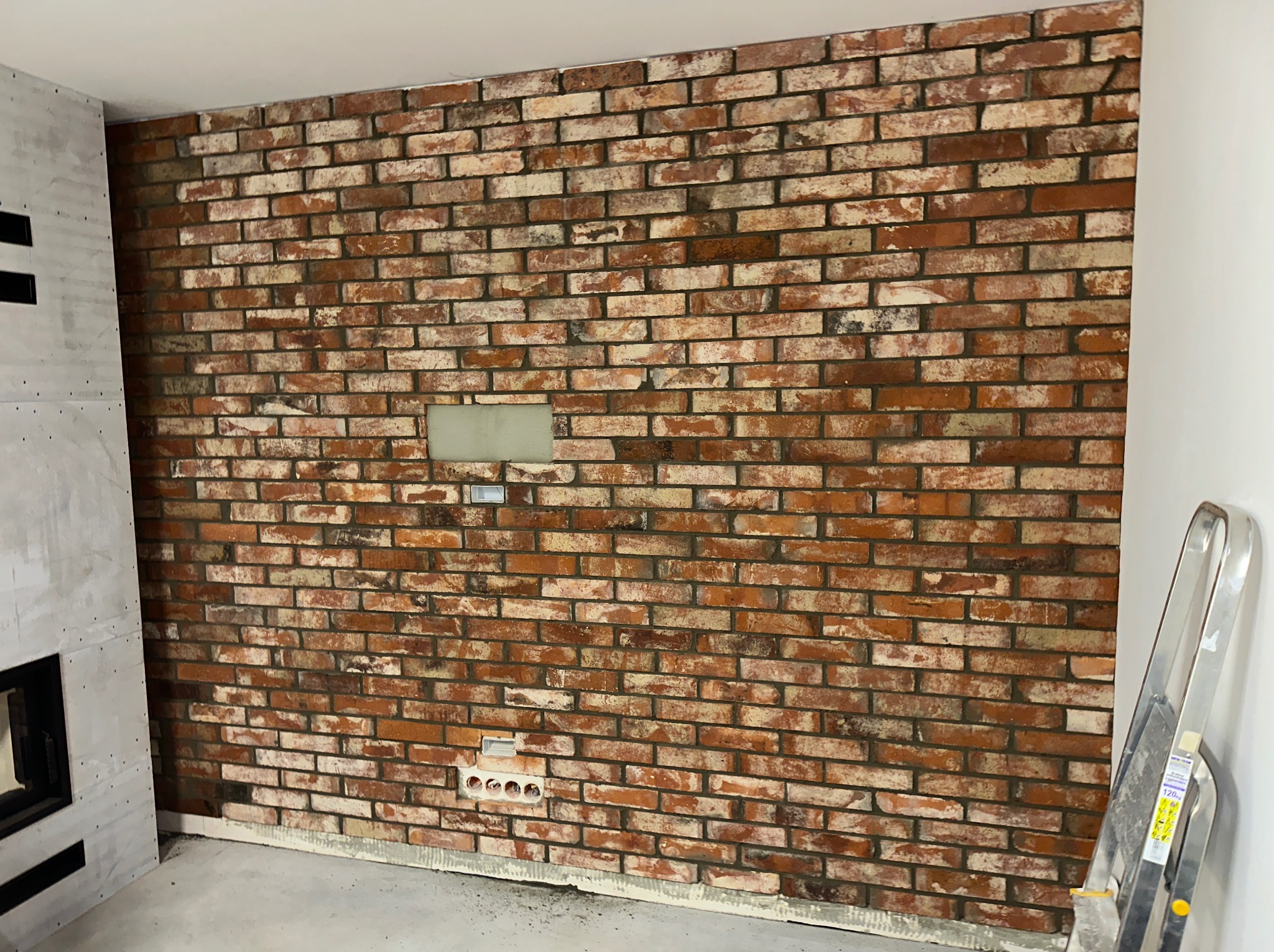Ściana z cegły klinkierowej w trakcie wykańczania, widoczne puszki elektryczne i częściowo wykończony kominek z płyt gipsowo-kartonowych.