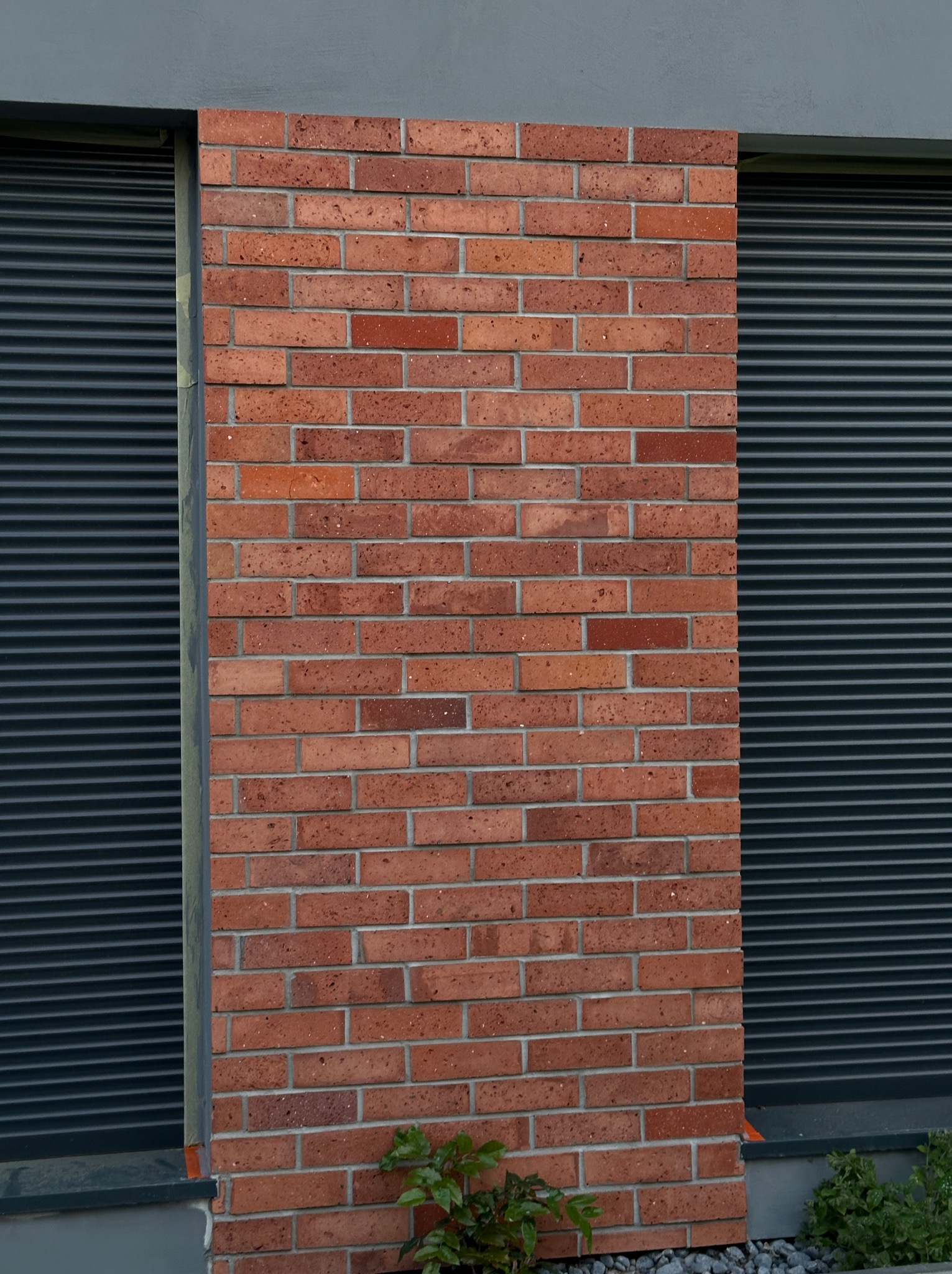 Fragment elewacji budynku z cegły klinkierowej w odcieniach czerwieni i brązu, przylegający do okna z szarymi roletami zewnętrznymi, z roślinami u podstawy ściany.