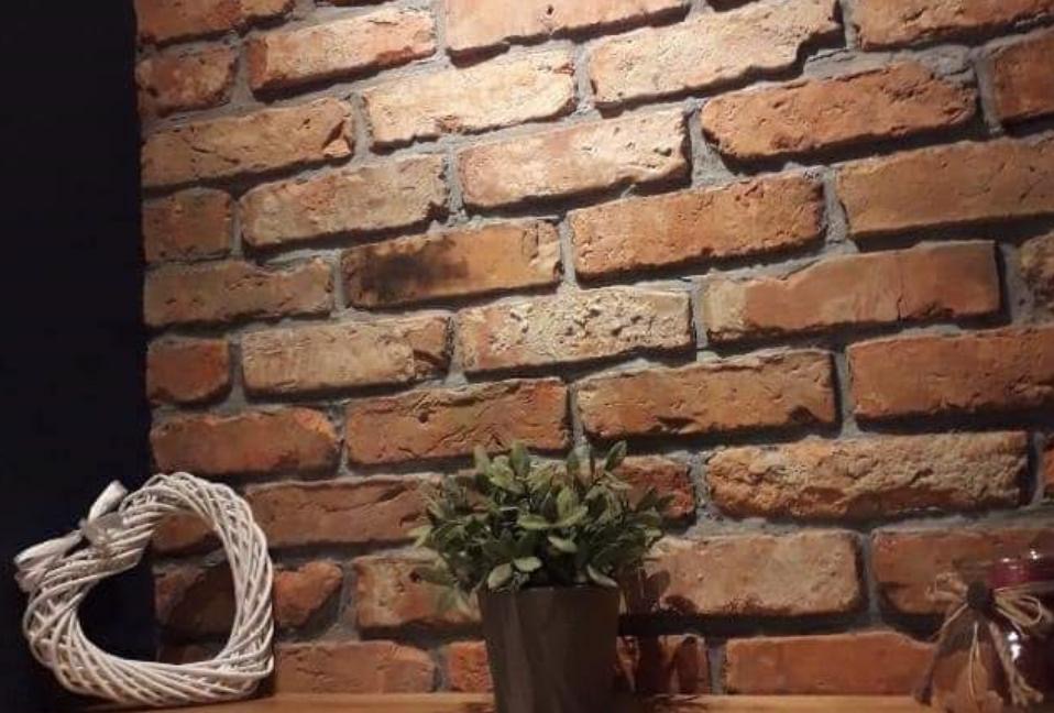 Dekoracyjna ściana z czerwonej cegły w Gnieźnie, z wiklinowym sercem i małą rośliną doniczkową na drewnianej półce.