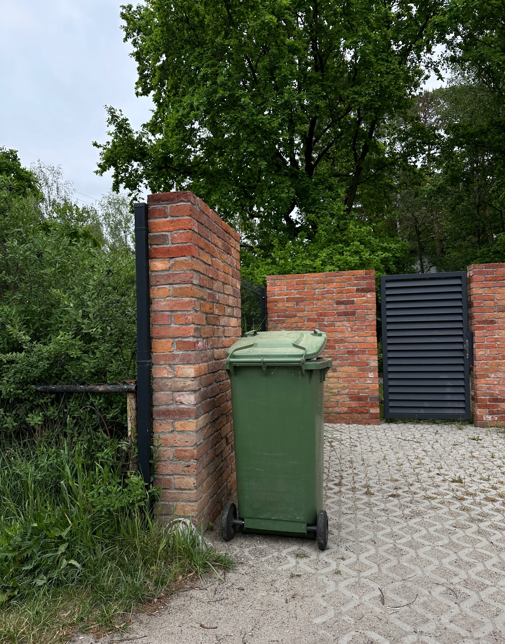 Ceglany mur z furtką i czarną, metalową bramą w tle, z zielonym pojemnikiem na odpady na pierwszym planie, stojącym na betonowej kostce.