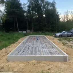 Przygotowany szalunek fundamentowy z ułożonym zbrojeniem siatkowym, widoczne pionowe rury instalacyjne, teren wokół wyrównany piaskiem, w tle zieleń i zaparkowany samochód.