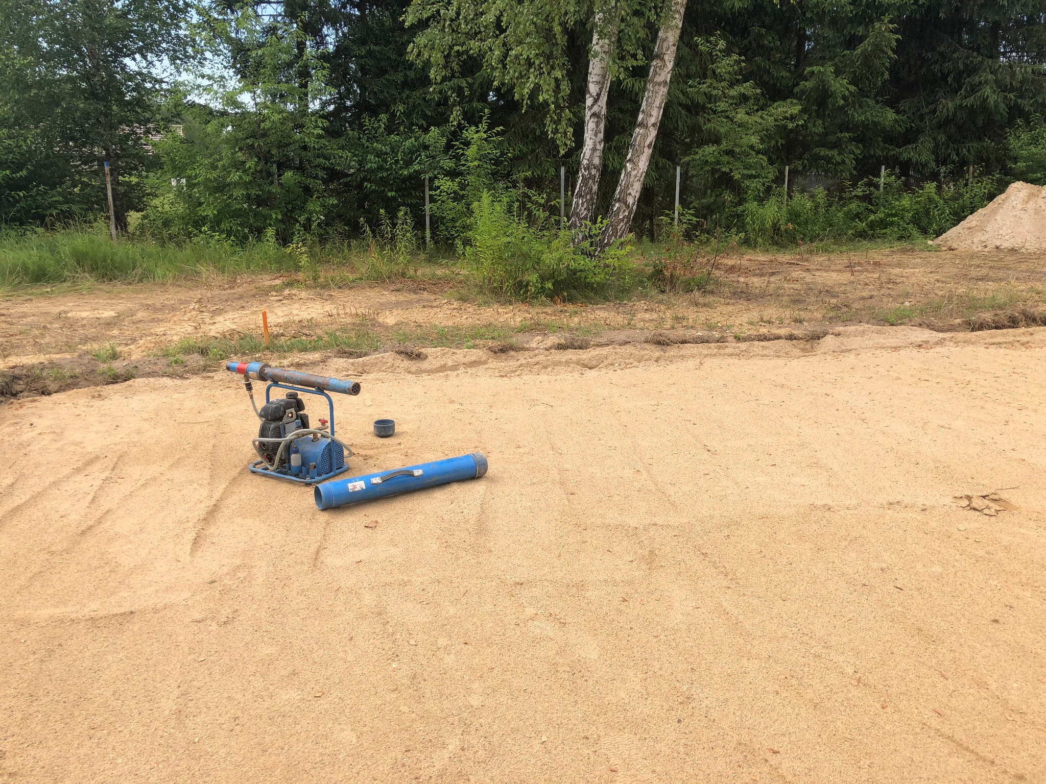 Wyrównany teren pod budowę z ubijarką wibracyjną i stertą piasku w tle, otoczony zielenią.