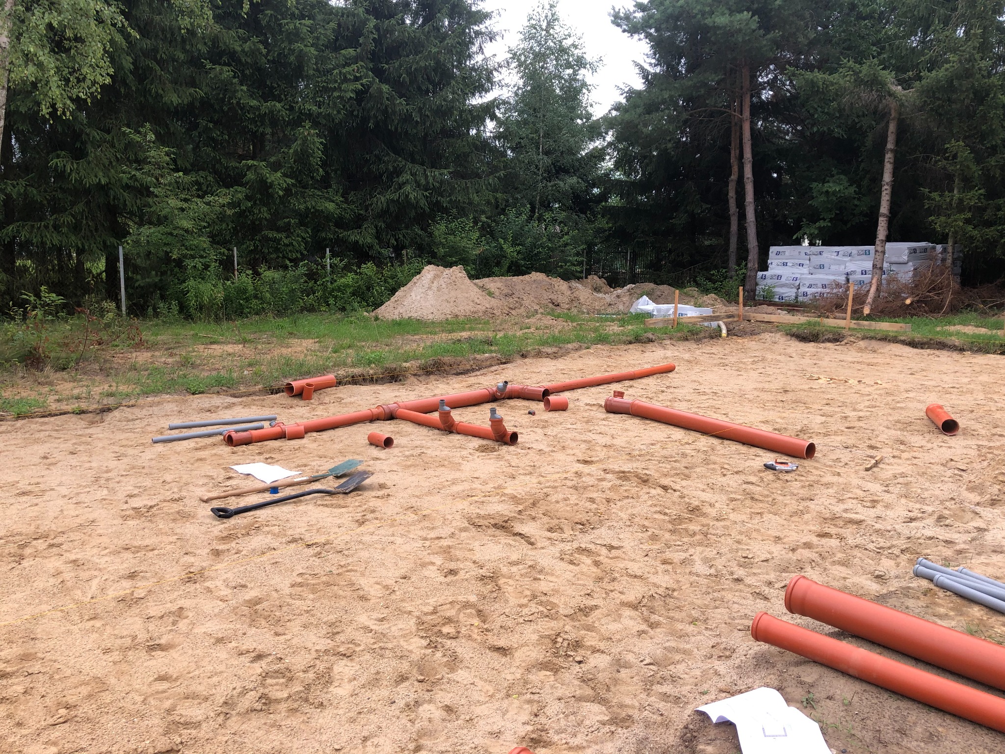 Przygotowanie terenu pod fundamenty: rozłożone pomarańczowe rury kanalizacyjne na piaszczystym gruncie, widoczne szpadle i dokumentacja, w tle stos materiałów budowlanych i drzewa.