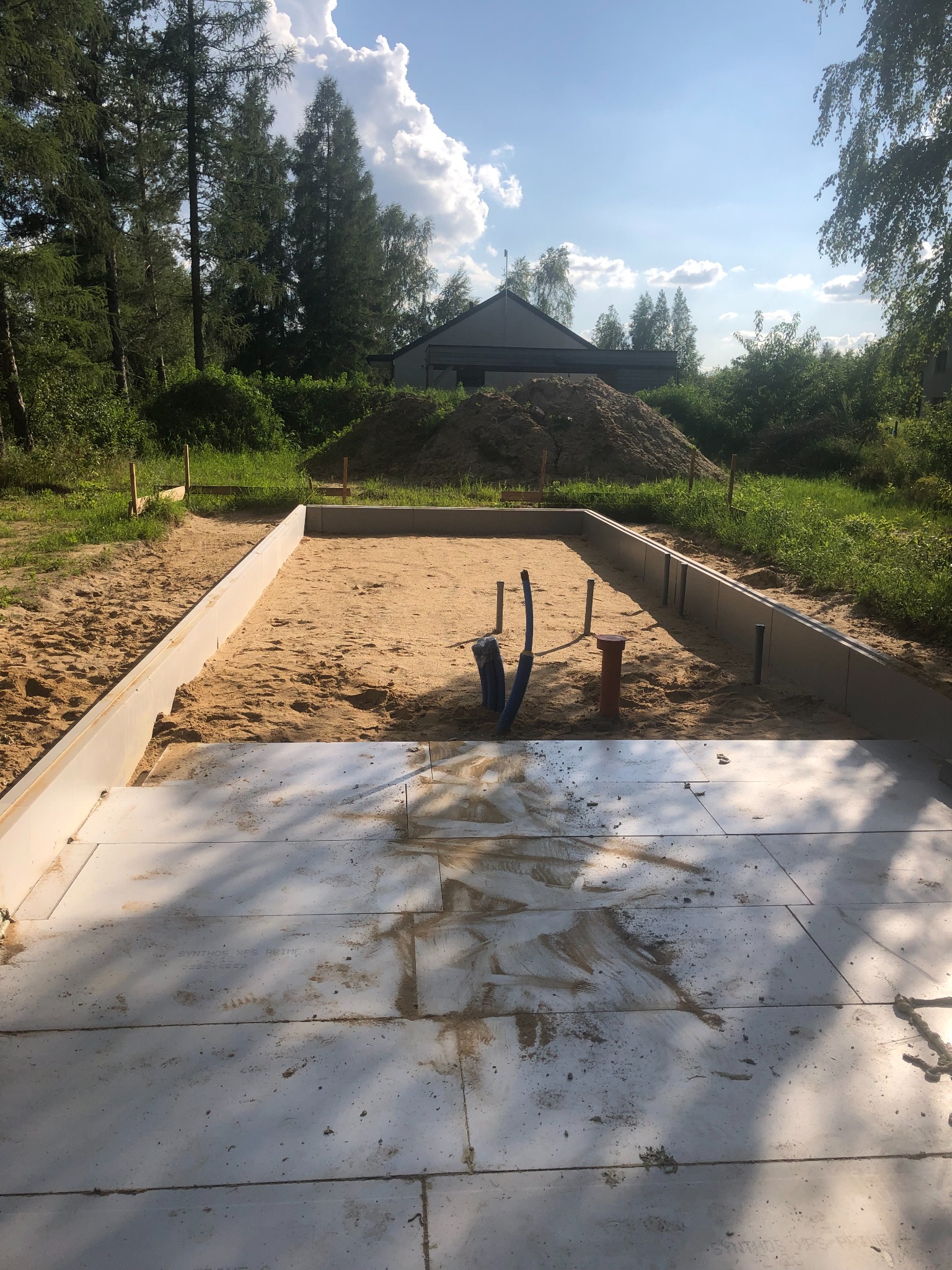 Przygotowany teren pod budowę domu: fundamenty szalunkowe, piasek, płyty izolacyjne, wystające rury instalacji wodno-kanalizacyjnej, w tle sterta ziemi i zabudowania gospodarcze.