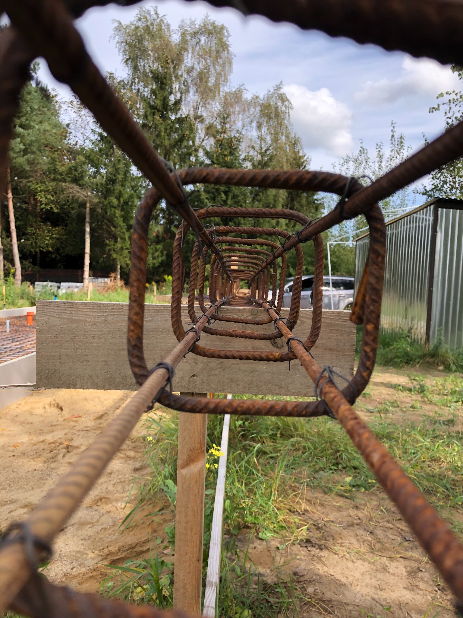 Zbrojenie fundamentowe ułożone w kwadratowy kształt, widok z perspektywy tunelu, z drewnianym podparciem i widocznym terenem budowy w tle.