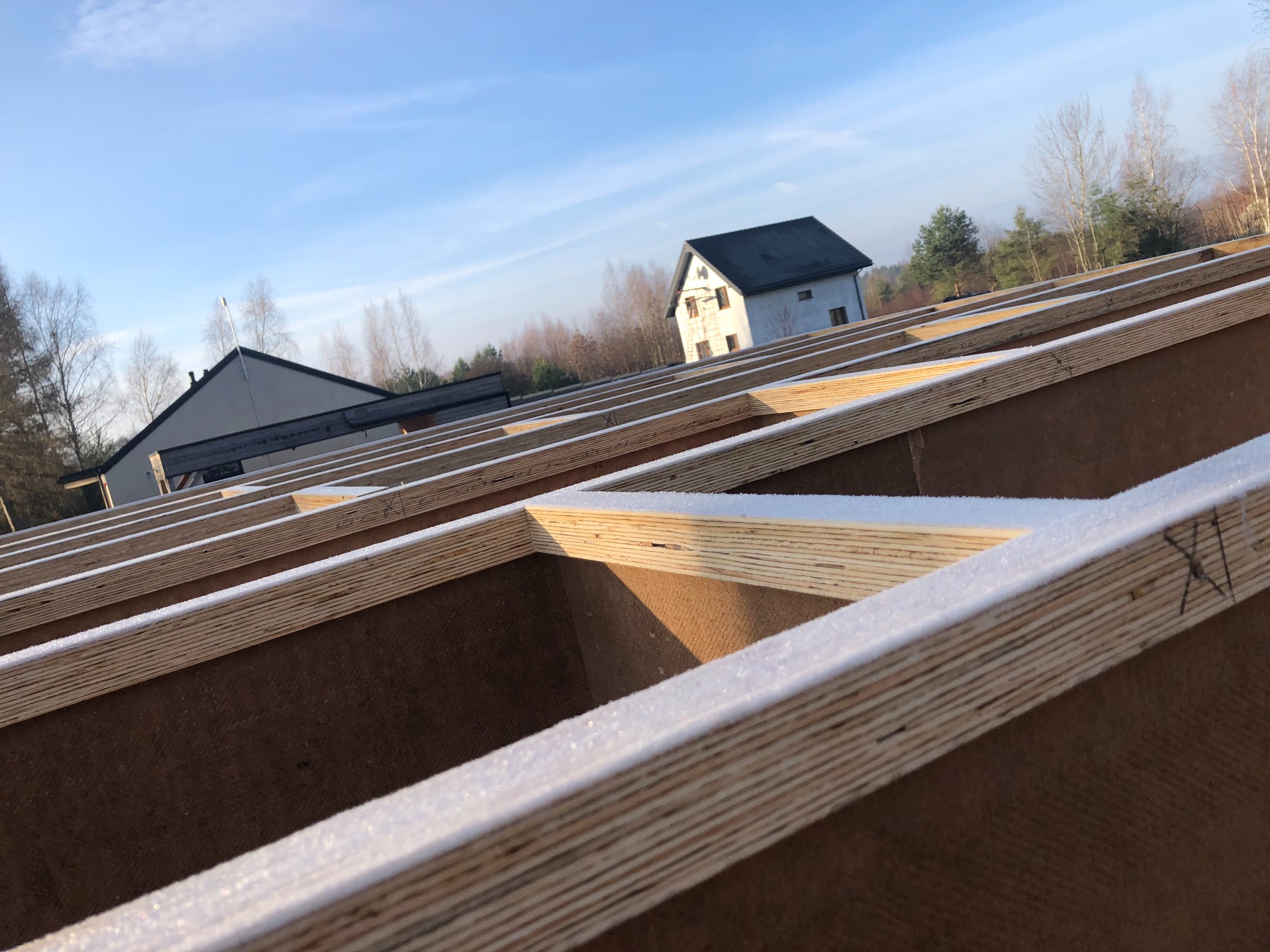 Wczesnym rankiem w Warszawie, widok na konstrukcję więźby dachowej pokrytej szronem, z dwoma budynkami w tle pod błękitnym niebem.