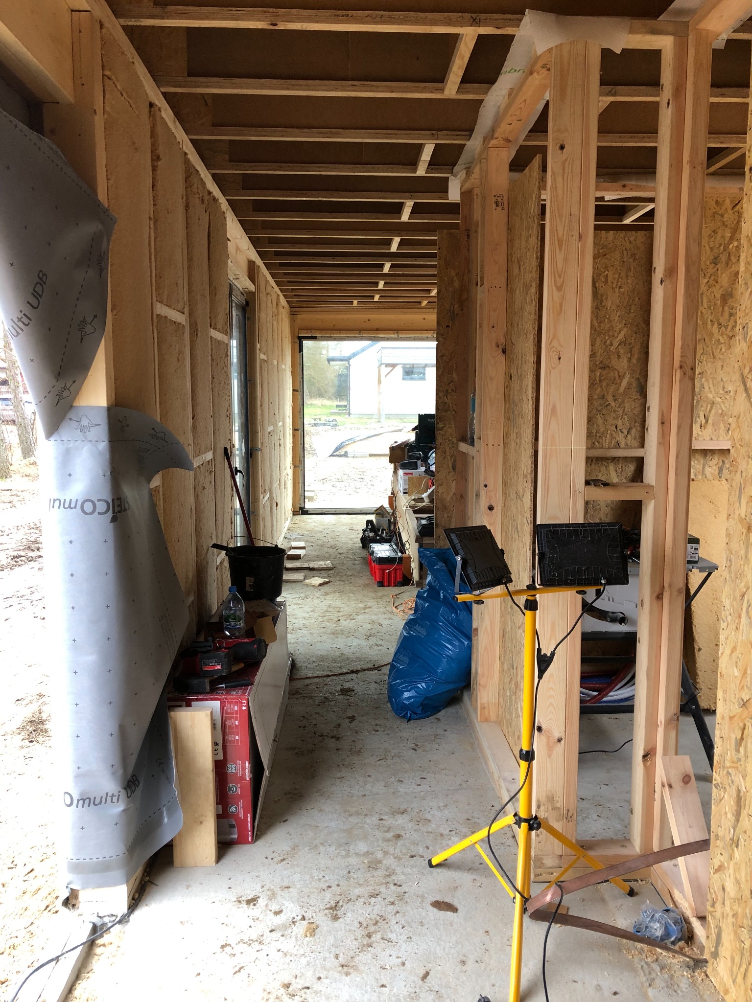 Wnętrze w trakcie budowy: surowe drewniane ramy ścian, izolacja, folia budowlana, tymczasowe oświetlenie robocze, widok na zewnątrz przez otwór okienny.