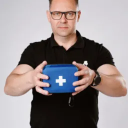 Mężczyzna w okularach i czarnej koszulce polo prezentuje niebieską apteczkę pierwszej pomocy z białym krzyżem na jasnoszarym tle.