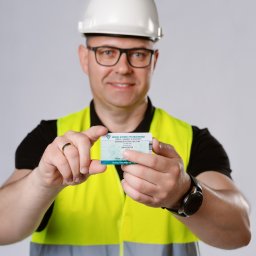 Inspektorek.pl Łukasz Jedliński - Uśmiechnięty mężczyzna w okularach, kasku i kamizelce odblaskowej prezentuje kartę kwalifikacyjną Urzędu Dozoru Technicznego.