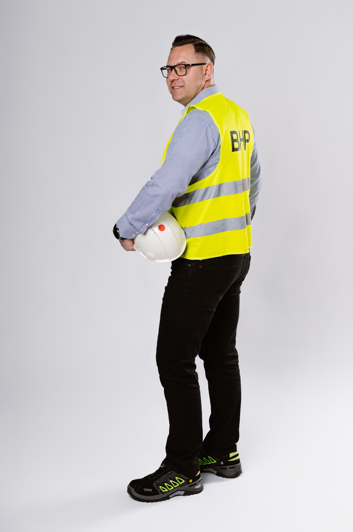 Uśmiechnięty mężczyzna w okularach, koszuli w paski i żółtej kamizelce z napisem BHP, trzymający biały kask ochronny, stojący na jasnym tle.