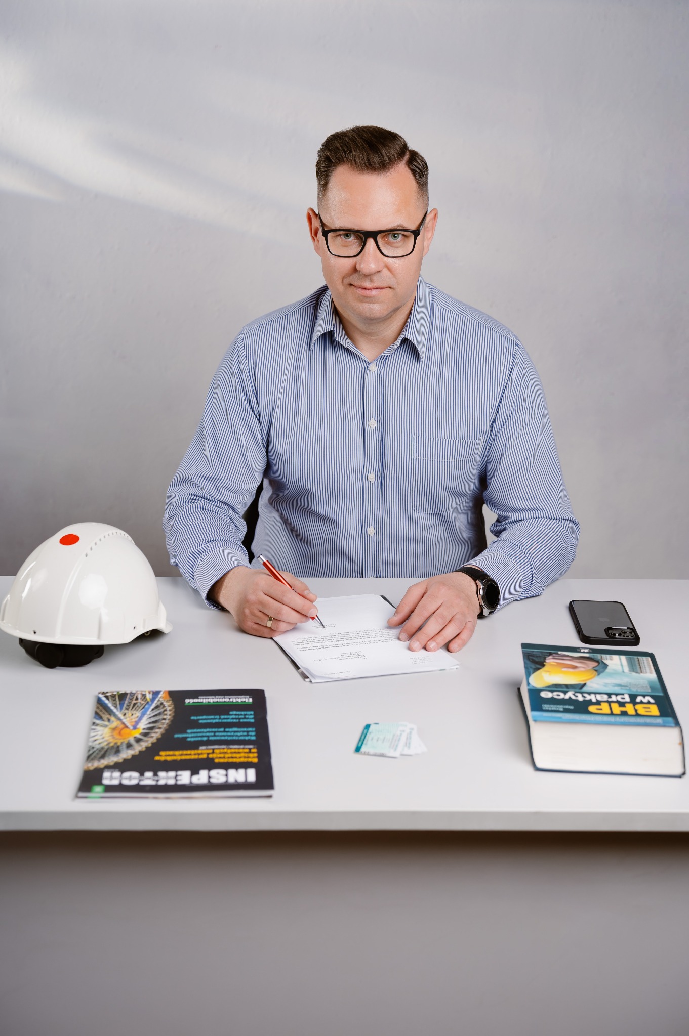 Mężczyzna w okularach i koszuli w paski siedzi przy biurku, podpisuje dokument, obok kask ochronny, telefon, książka 'BHP w praktyce' i magazyn 'Inspektor'.