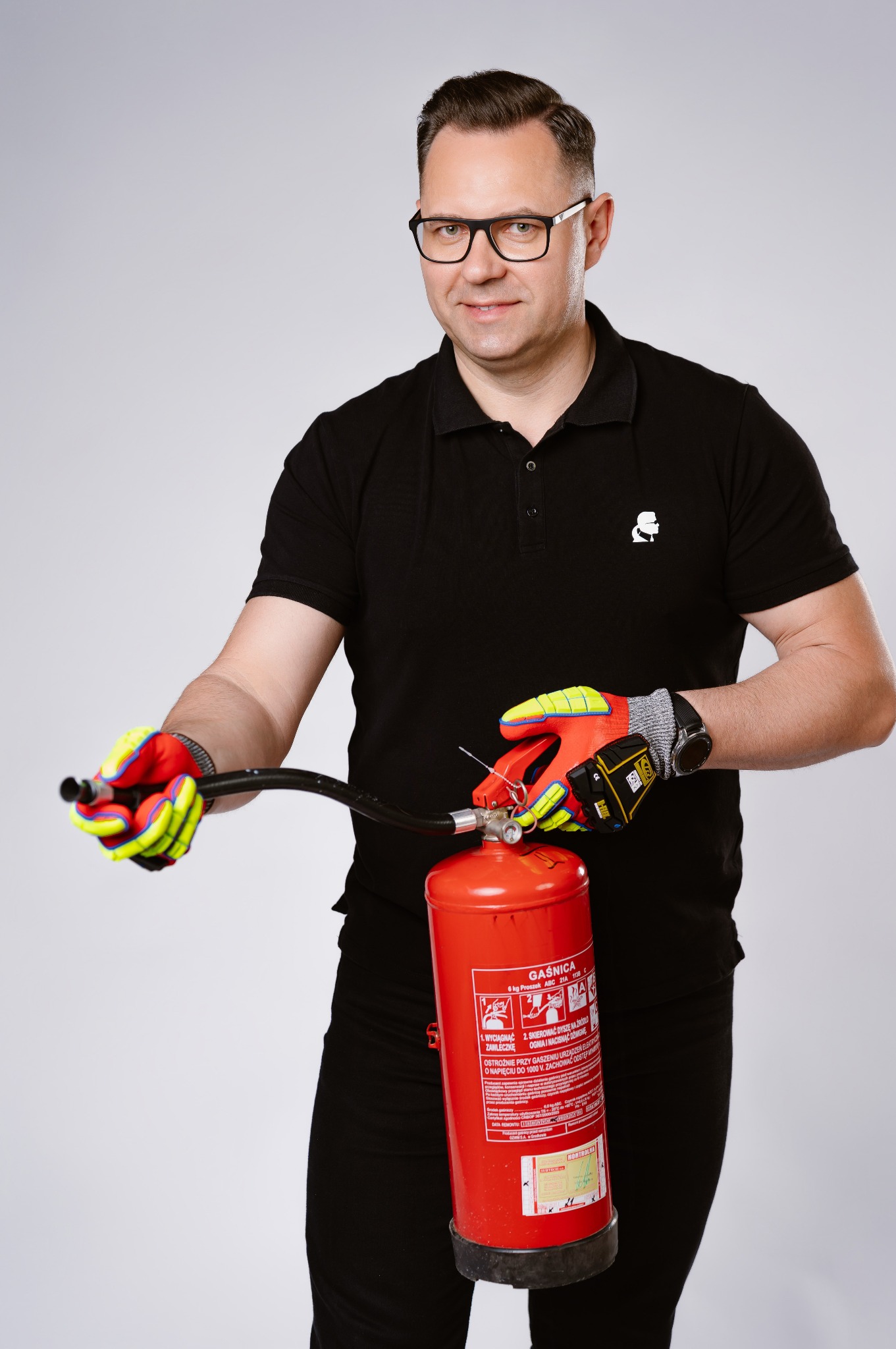 Mężczyzna w okularach i czarnej koszulce polo trzyma czerwoną gaśnicę, ubrany w robocze rękawice, przygotowując się do użycia.