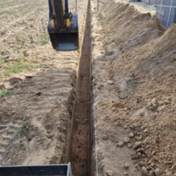 Żółto-czarna koparka pogłębia wąski, prosty wykop w piaszczystej glebie, z widocznymi stertami ziemi po bokach i napiętym sznurkiem wyznaczającym linię, w tle budynek w trakcie budowy i pole uprawne.