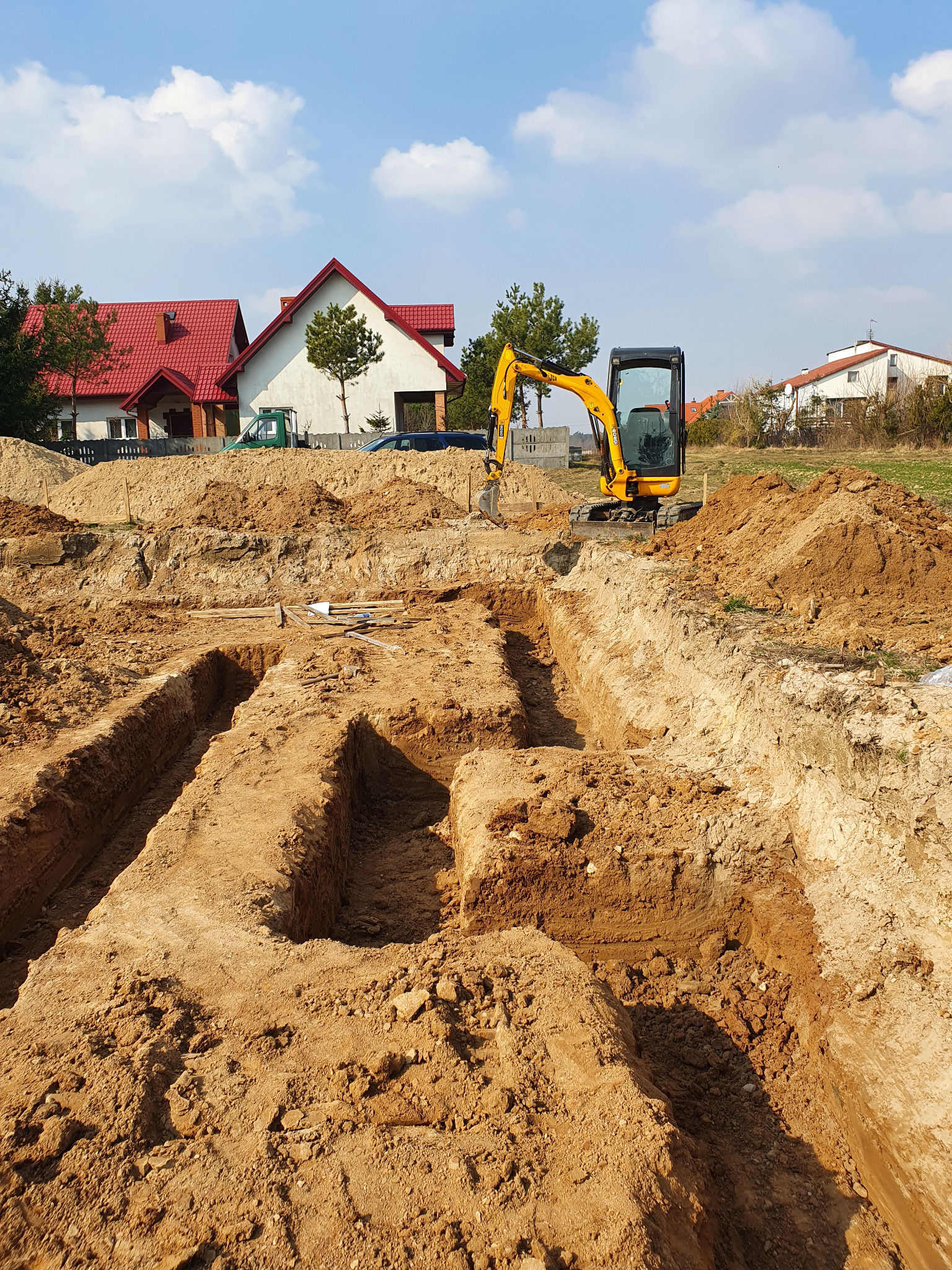 Żółta mini koparka JCB stoi na placu budowy obok wykopanych rowów pod fundamenty, w tle domy jednorodzinne pod błękitnym niebem.