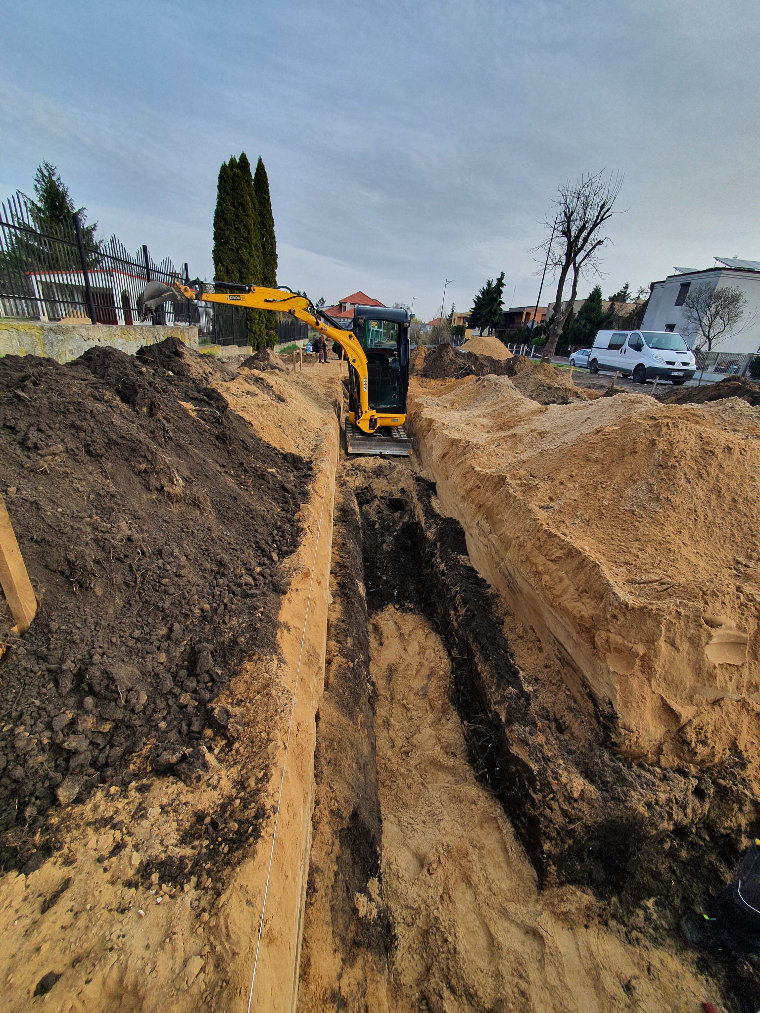 Żółta mini koparka w trakcie wykonywania głębokiego wykopu w piaszczystej glebie, widoczne warstwy ziemi i ułożone pryzmy ziemi po bokach wykopu, w tle zabudowa jednorodzinna i pochmurne niebo.
