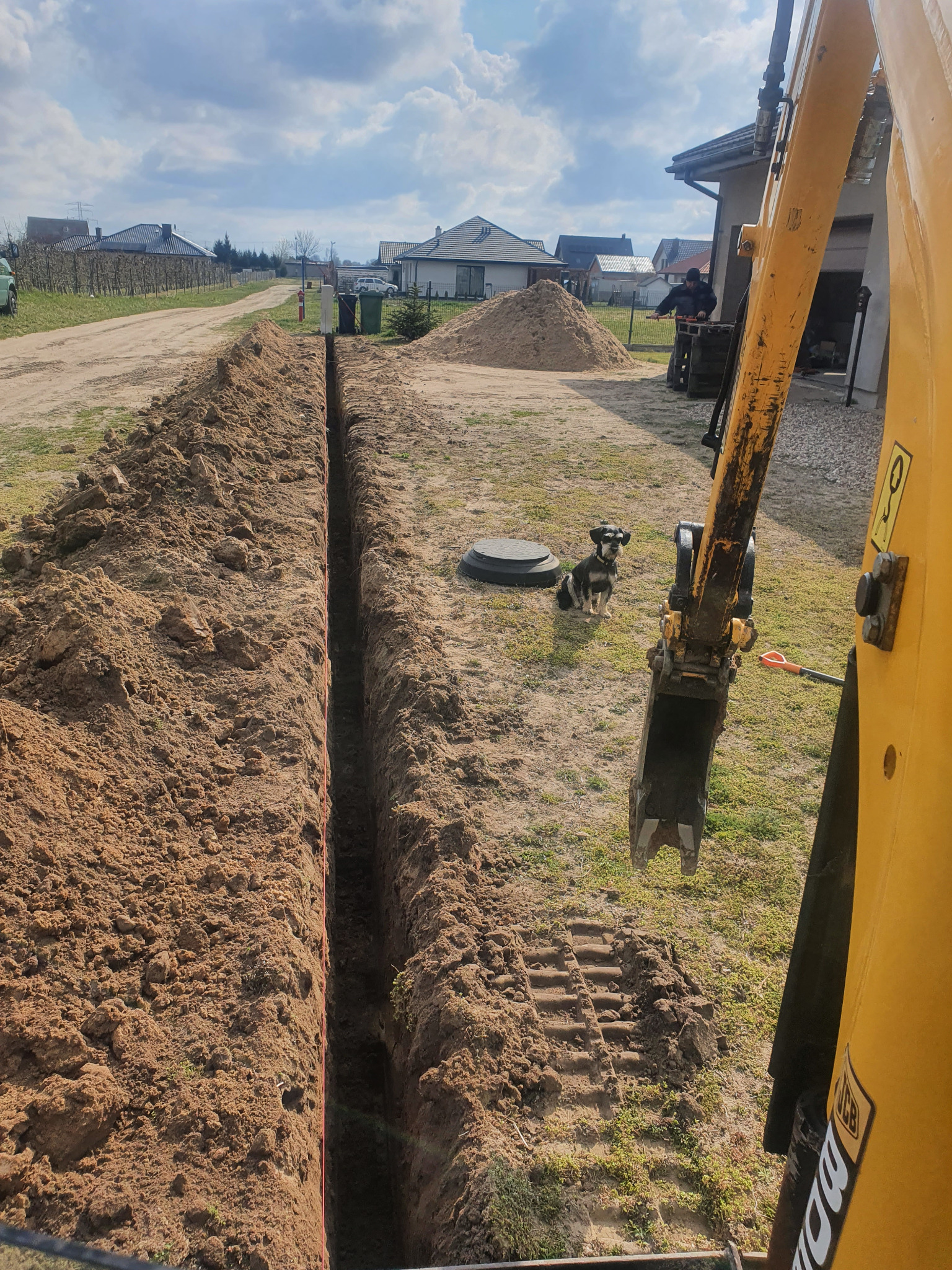 Wąski wykop w ziemi, wyznaczony sznurkiem, z widoczną koparką JCB i psem rasy sznaucer miniaturowy w tle, na działce z zabudową jednorodzinną.