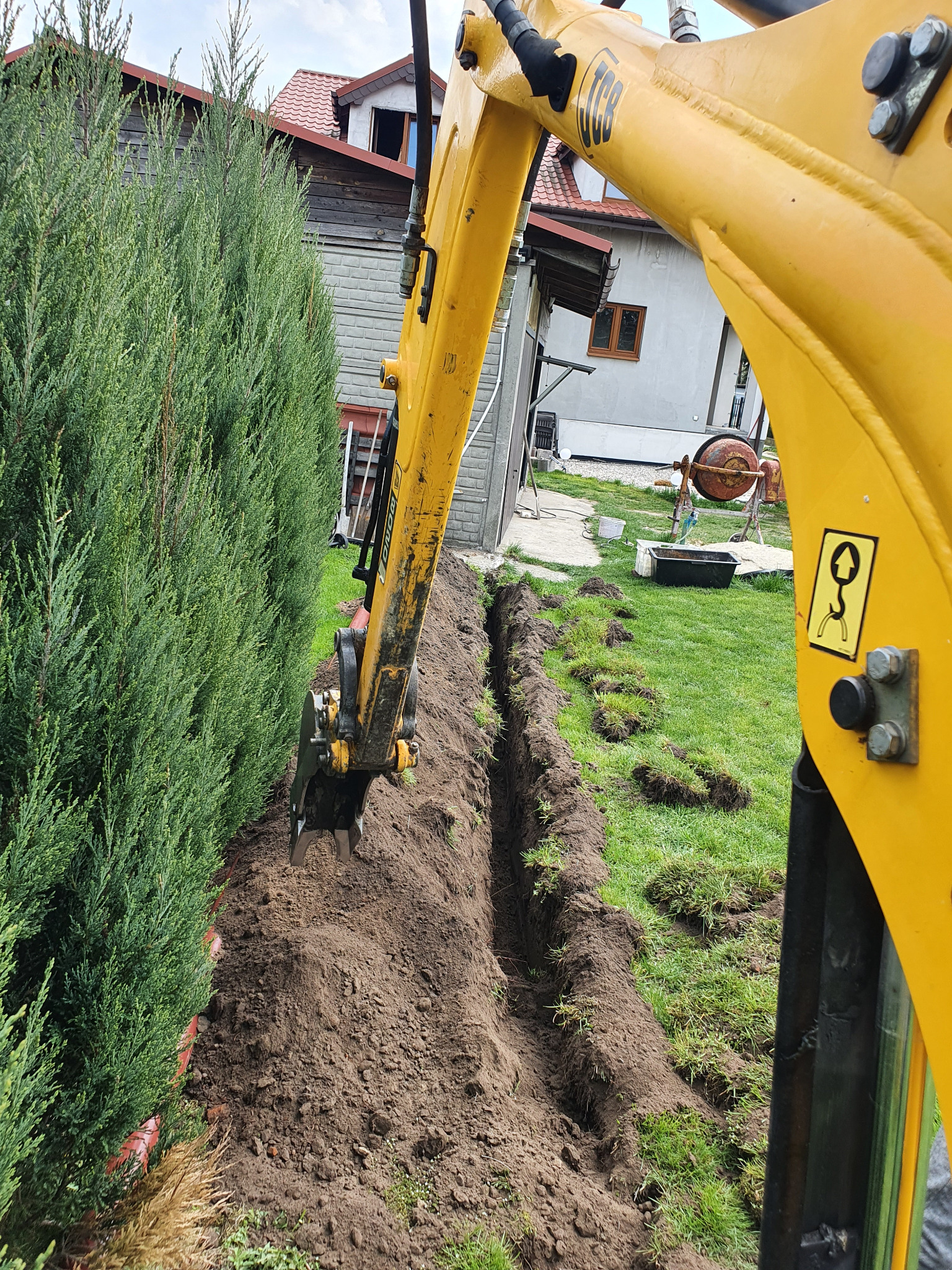 Żółta koparka JCB w trakcie wykonywania wąskiego wykopu w ziemi, widoczne świeżo wykopane fragmenty trawy, w tle dom jednorodzinny i wysokie tuje.