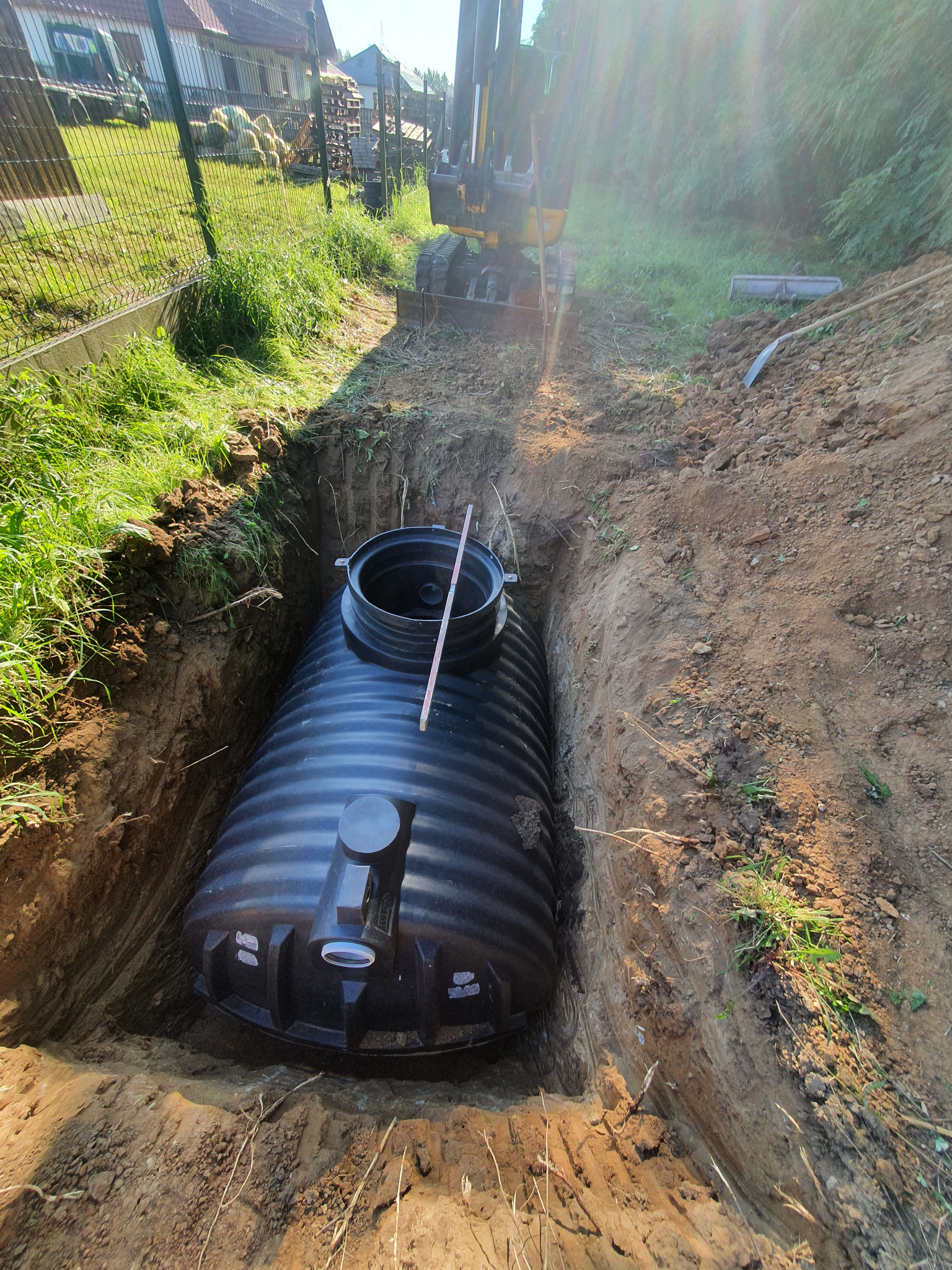 Czarny, żebrowany zbiornik na szambo umieszczony w wykopie z żółtą ziemią, z tyłu widoczna koparka i zielona trawa.