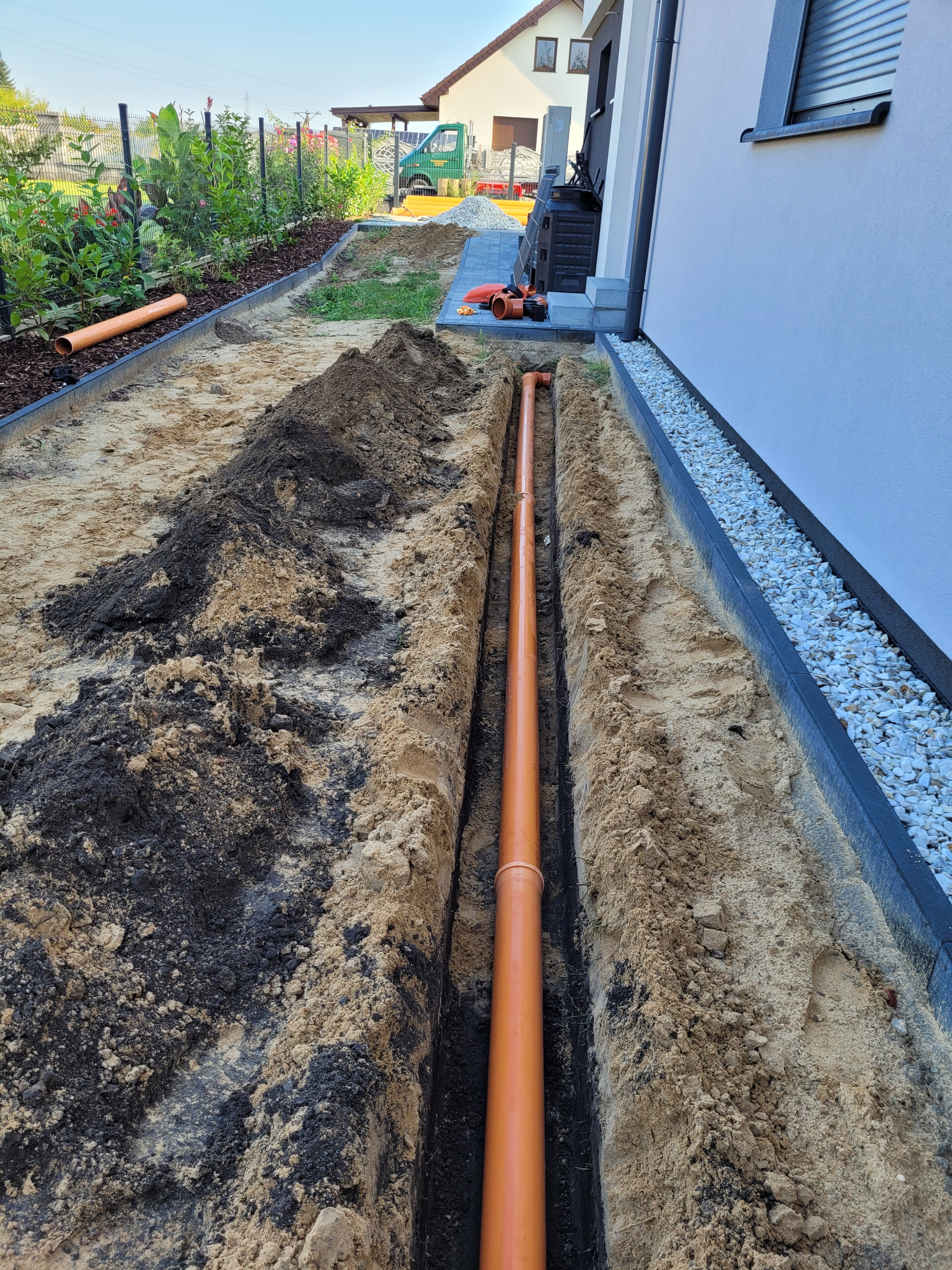 Pomarańczowa rura ułożona w wykopie wzdłuż ściany budynku, widoczny piasek, czarnoziem i obrzeża trawnika.
